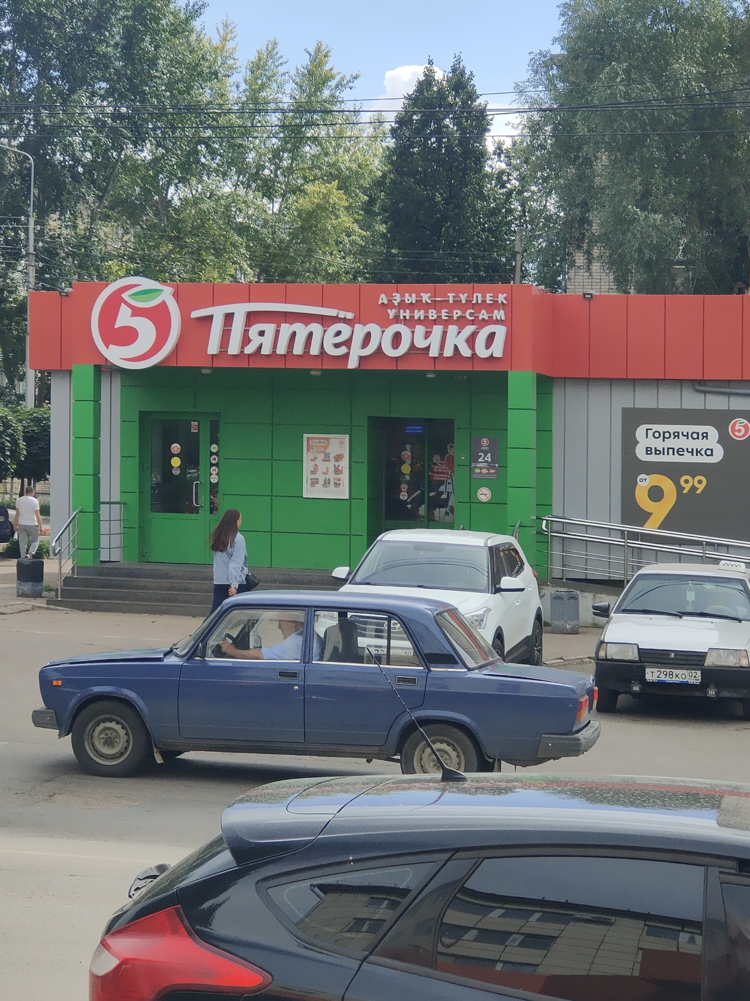 In Oktyabrsky, Russia, a "Pyaterochka" supermarket with a bright green facade is visible, displaying its signature red and white logo. Above the green section, a red awning runs the width of the building, also featuring the "Pyaterochka" branding. To the right of the entrance, a black sign advertises "Hot baked goods" with a price of 99. A woman in a denim jacket stands on the sidewalk, looking towards the entrance, while a man in a white t-shirt walks away from the building. Several cars are parked on the street, including a blue sedan and two white cars, one of which is a white Lada. The street is paved, and a sidewalk with steps leads up to the supermarket entrance. Trees line the background, suggesting an urban environment. A black car is parked in the foreground, partially obscuring the view of the other vehicles and the street.