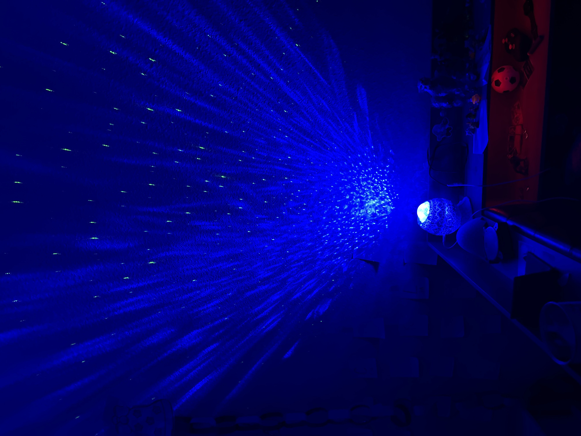 An indoor scene, likely a bedroom or playroom, is illuminated by a blue projector creating a starry night sky effect on the ceiling and walls. The projector itself is a small, decorative lamp with a textured, cracked surface, emitting a bright blue light that spreads outward in radiating beams. Scattered across the ceiling and walls are small, faint green pinpoints of light, adding to the celestial illusion.
In the lower part of the image, a dark wooden shelf or desk is visible, upon which several objects are placed. These include a small soccer ball with black and white panels, a dark-colored game controller, and other indistinct toys or figurines that are mostly obscured by shadows. A white cylindrical object, possibly a can or container, is also partially visible on the left side of the shelf. The overall ambiance is dark and somewhat whimsical, suggesting a child's private space. The image does not contain enough information to determine a specific location within Colorado Springs, United States of America.