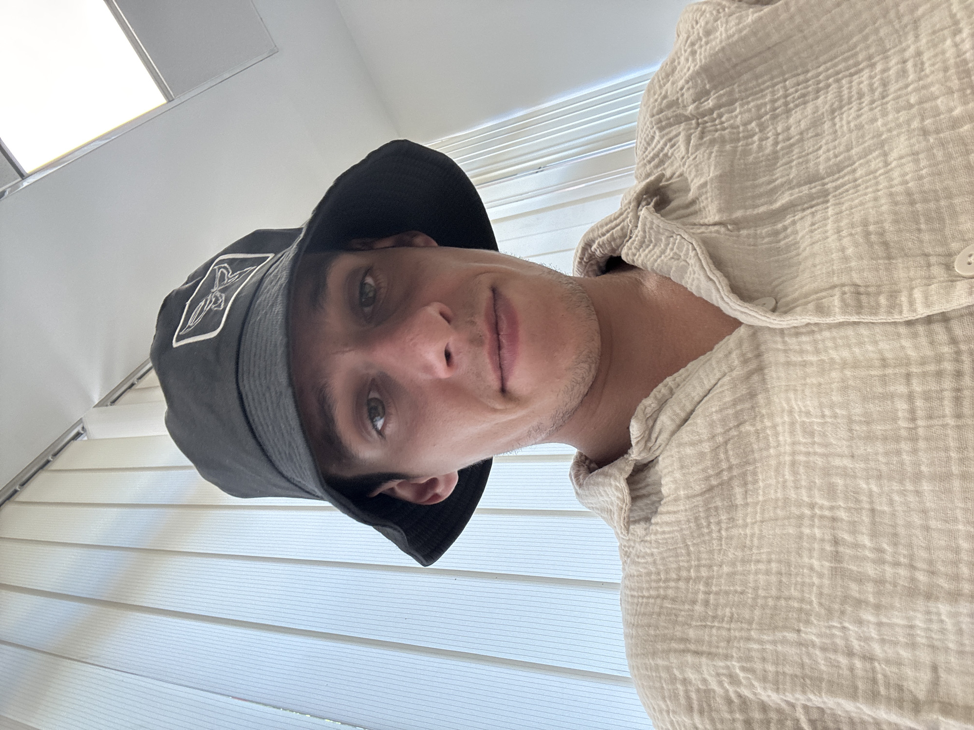 The image shows a close-up, front-facing selfie of a young adult male wearing a black bucket hat and a light-colored, textured button-up shirt. He has short brown hair visible beneath the hat, and his eyes are looking directly at the camera. The background consists of vertical blinds and a ceiling light fixture. The overall setting appears to be indoors, possibly an office or a room with vertical window coverings. There is no indication of the location being in Sofia, Bulgaria.
