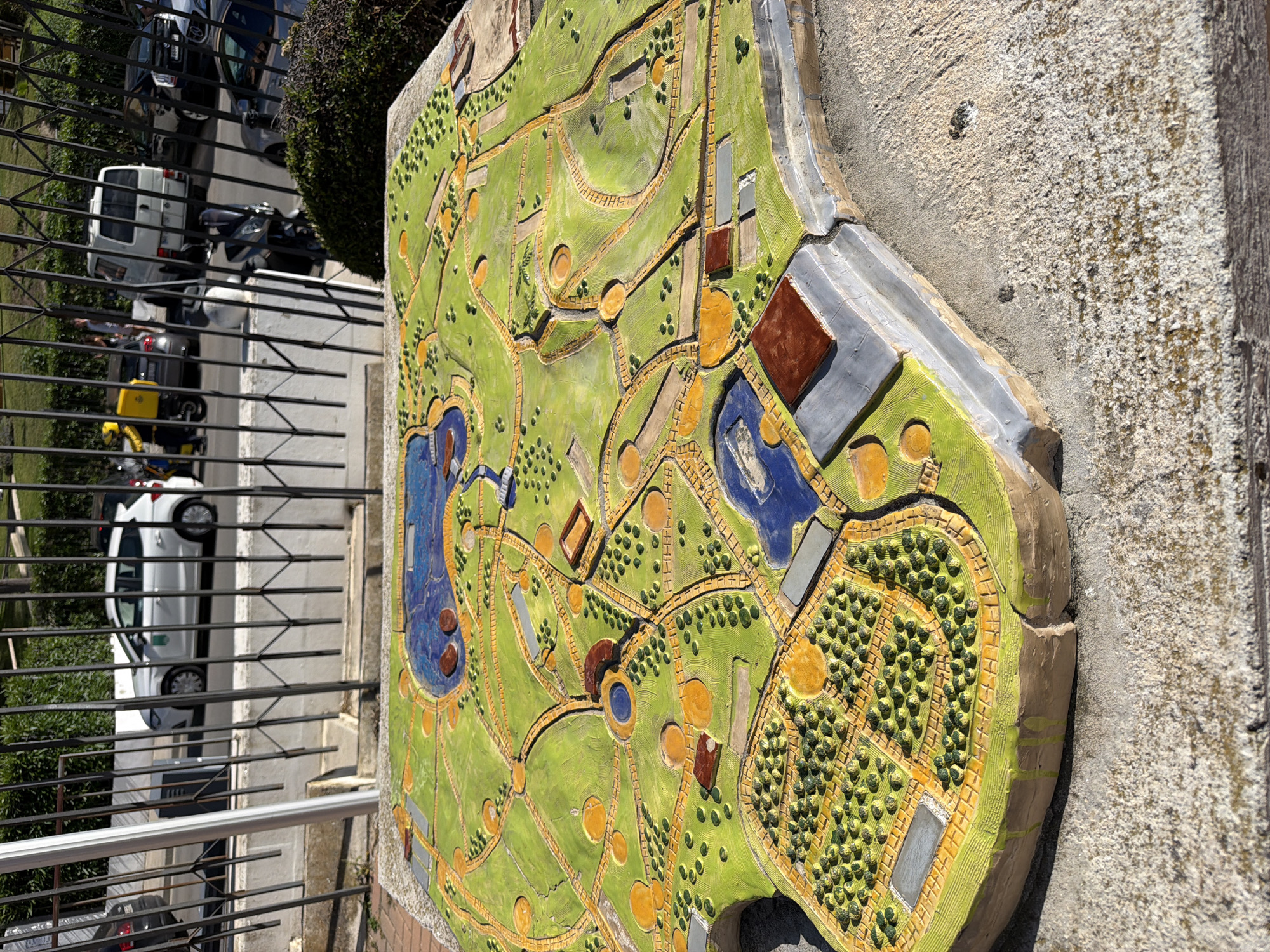 A high-angle, slightly tilted shot presents a park map in Arroyo De La Miel-Benalmadena Costa, Spain, mounted on a wall behind a black metal fence. The map, made of textured materials like paint and possibly tile, depicts a green park landscape with winding yellow paths, representations of bodies of water (blue), tree clusters (green dots), buildings (brown and grey rectangles), and circular areas. The surface on which the map is mounted is textured concrete. Outside the fence, a view of a street includes parked cars, a small green bush, and parts of buildings. The primary subject is the park map itself. The day appears sunny, casting shadows and highlighting the map's texture.