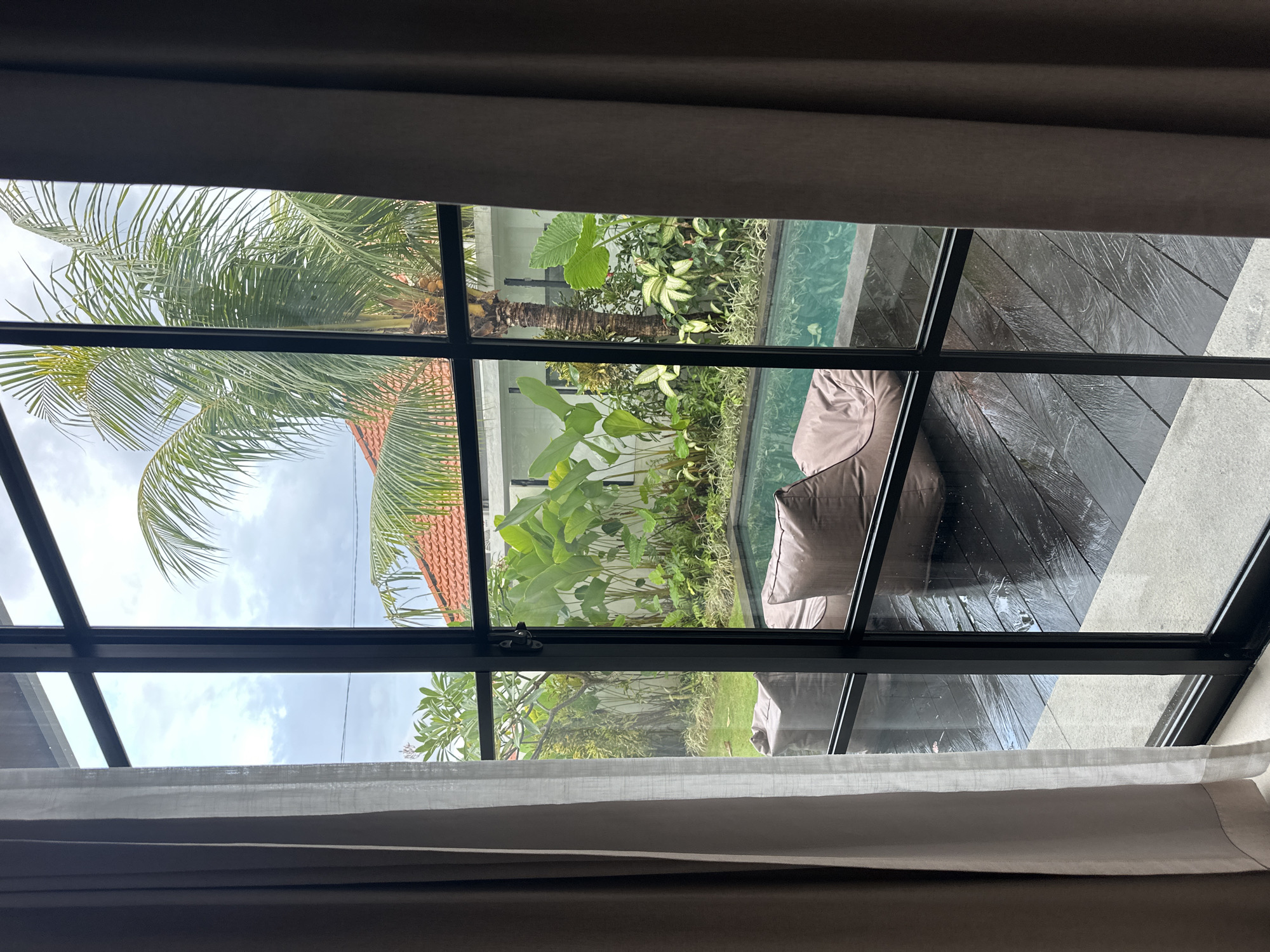 The image captures an indoor scene looking out through a multi-pane window towards an outdoor area. The window is framed by gray curtains, partially drawn, revealing a view of a lush tropical garden. Outside, a palm tree and various green plants thrive, suggesting a warm, humid climate. A section of a red-tiled roof is visible, indicating nearby buildings. The garden also includes a small swimming pool, adjacent to a dark wooden deck where what appear to be tan beanbag chairs are placed. The overall setting suggests a private villa or residence in a tropical location. The location is identified as North Kuta, Indonesia. The weather appears cloudy or overcast. There are no people visible in the image.