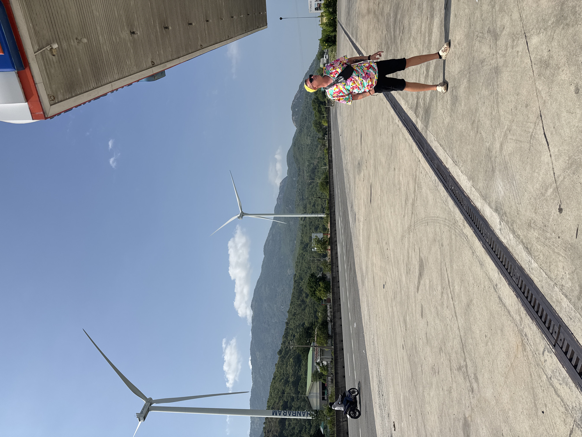 A man stands on a concrete lot under a gas station canopy. He wears a yellow hat, sunglasses, a brightly colored t-shirt, black shorts, and tan shoes. A black fanny pack is slung across his chest. He is looking up and to the right. In the background, a highway runs past a line of trees and hills. Two wind turbines are visible on the horizon. A motorbike is driving on the highway. The sky is clear and blue.