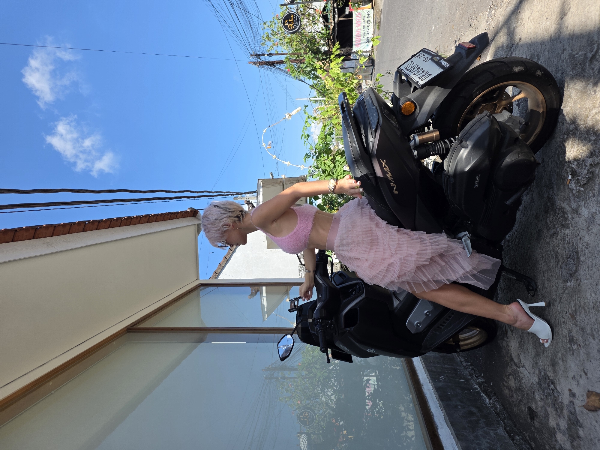 A woman with short, light-colored hair and sunglasses is sitting on a black Yamaha NMAX scooter parked on a paved road in front of a building with large glass windows and a tiled roof. She is wearing a pink fuzzy crop top and a tiered pink tulle skirt, along with white heeled mules. Her left hand is resting on the scooter's handlebar, and her right hand is holding a smartphone. In the background, there are power lines, lush green trees, and buildings. A sign in the background reads "Ratna Restaurant & Bar." The license plate on the scooter is DK 4649 FDL. The sky is blue with scattered white clouds.