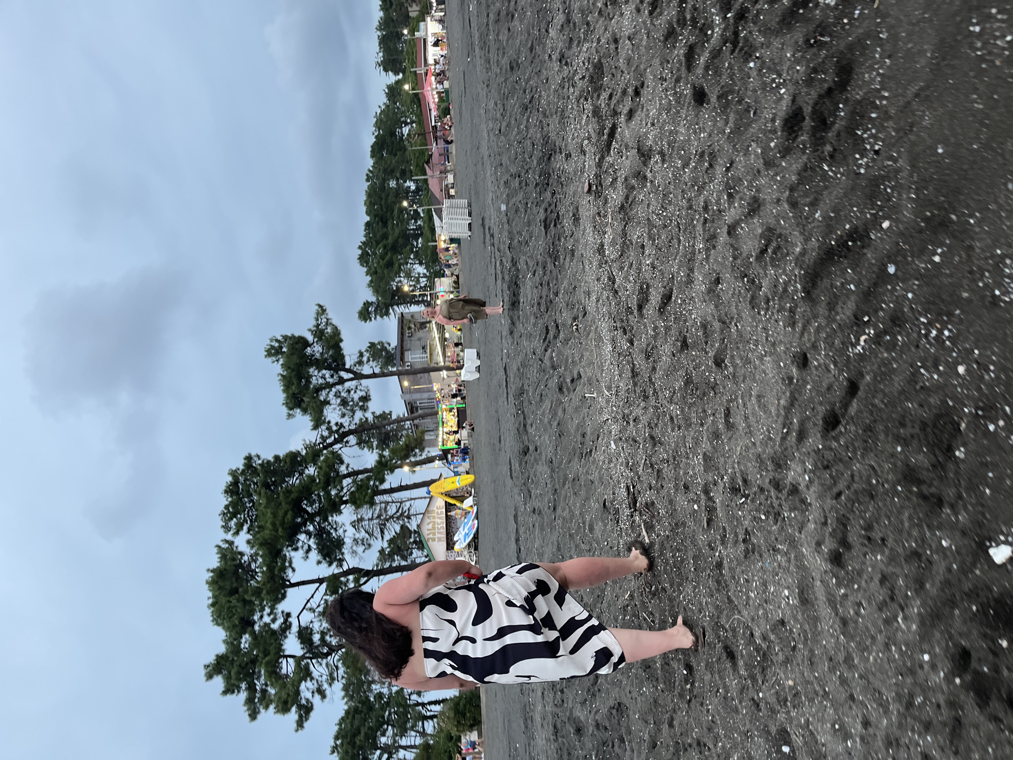 The image captures a scene on the black sand beach of Ureki, Georgia. In the foreground, a woman wearing a black and white patterned sarong walks away from the camera, her bare feet leaving imprints in the dark sand. Further back on the beach, another person in a green dress stands facing the water. The background reveals a lively stretch of beach with various vendors, tents, and people milling about, illuminated by ambient lighting. Pine trees line the edge of the beach, and the sky above is overcast with a pale, diffused light. Several white lounge chairs and surfboards are visible near the vendors, suggesting a typical beach day atmosphere. The overall environment is that of a public beach with commercial activity.