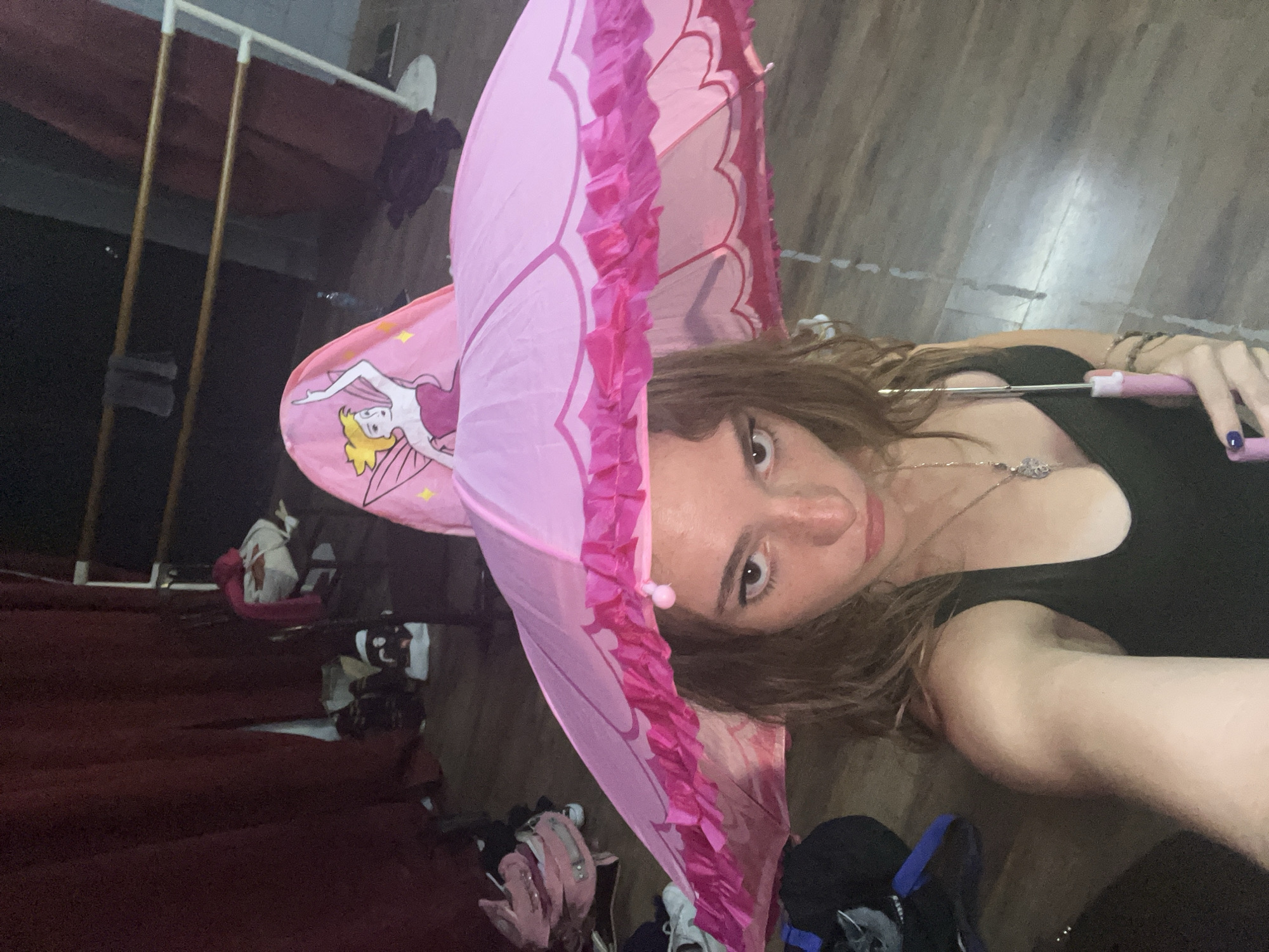 The image shows a woman indoors, holding a pink umbrella with a cartoon fairy illustration and ruffled edges. She is wearing a black tank top and a silver necklace. The background includes a red carpet, a clothes drying rack, and various personal belongings. The location is not evident from the image.