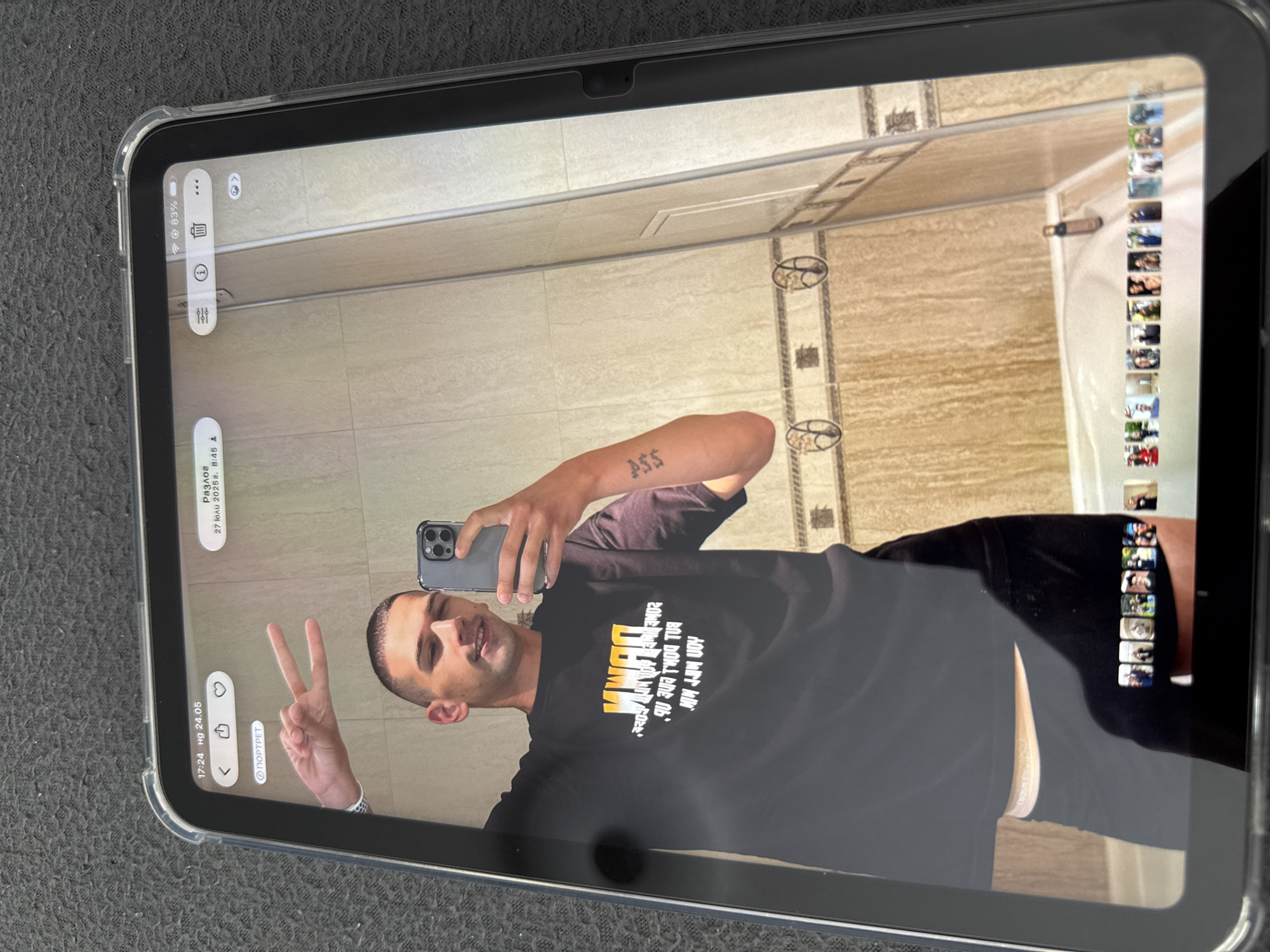 A man is taking a selfie in a bathroom using a phone. He is wearing a black t-shirt and black pants. He is making a peace sign with his left hand. The bathroom has light-colored tiles and a shower in the background. The tablet displaying the photo has Russian text and indicates the city of Razlog and a date of July 27, 2025. The battery level is at 83%.