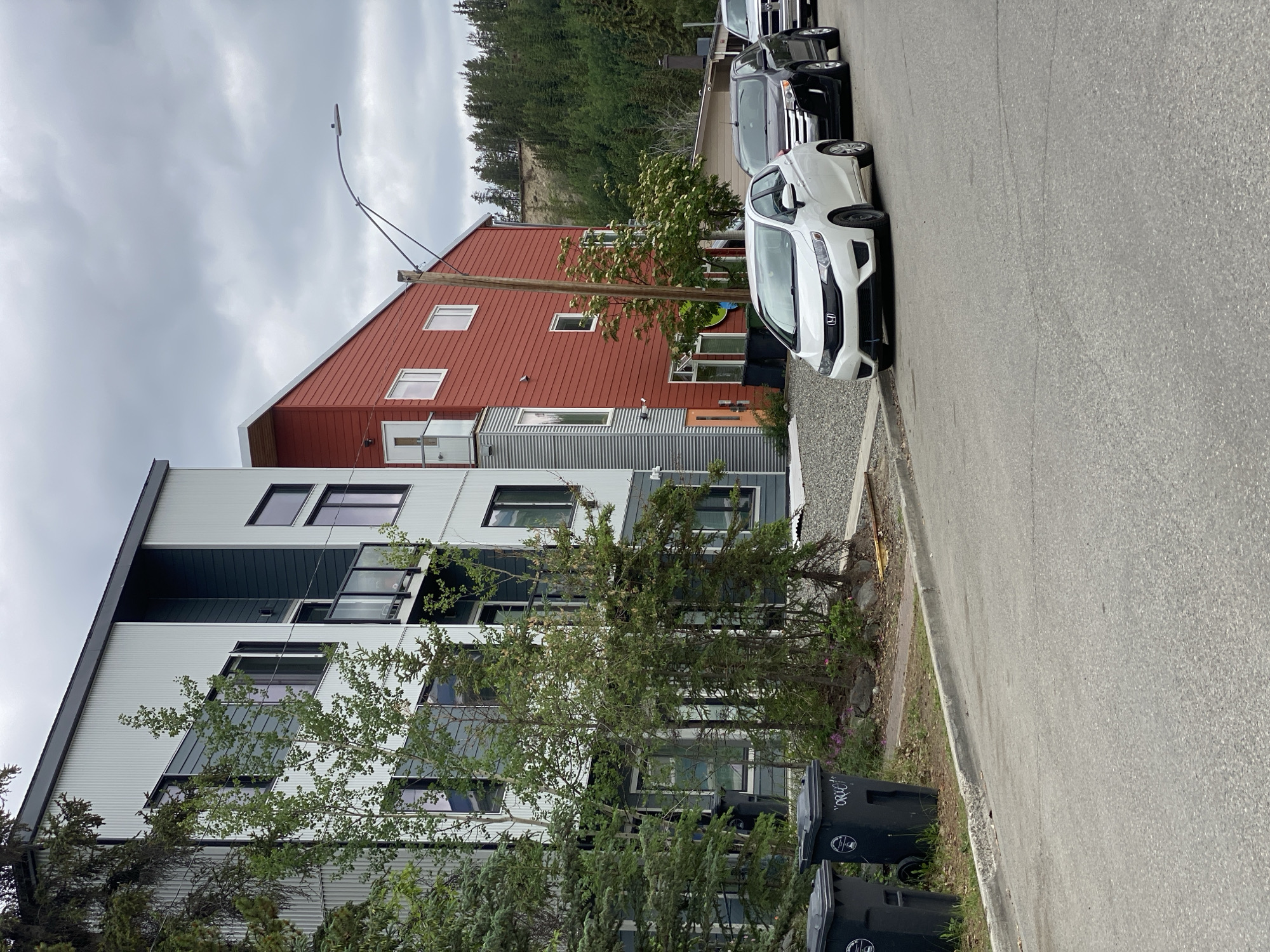 A residential street in Whitehorse, Canada, is depicted under an overcast sky. The image features a multi-story, modern building with a gray and teal facade on the left, and a striking red addition on the right. Trees are sparsely planted in front of the building, and two large black trash bins are visible on the left side of the image. A white Honda Fit is parked in front of the building, followed by a black SUV and a silver pickup truck. The street is paved, with a concrete curb and a gravel area bordering the building. In the background, a treed hillside rises, with some erosion visible at its crest. A street lamp arches over the scene.