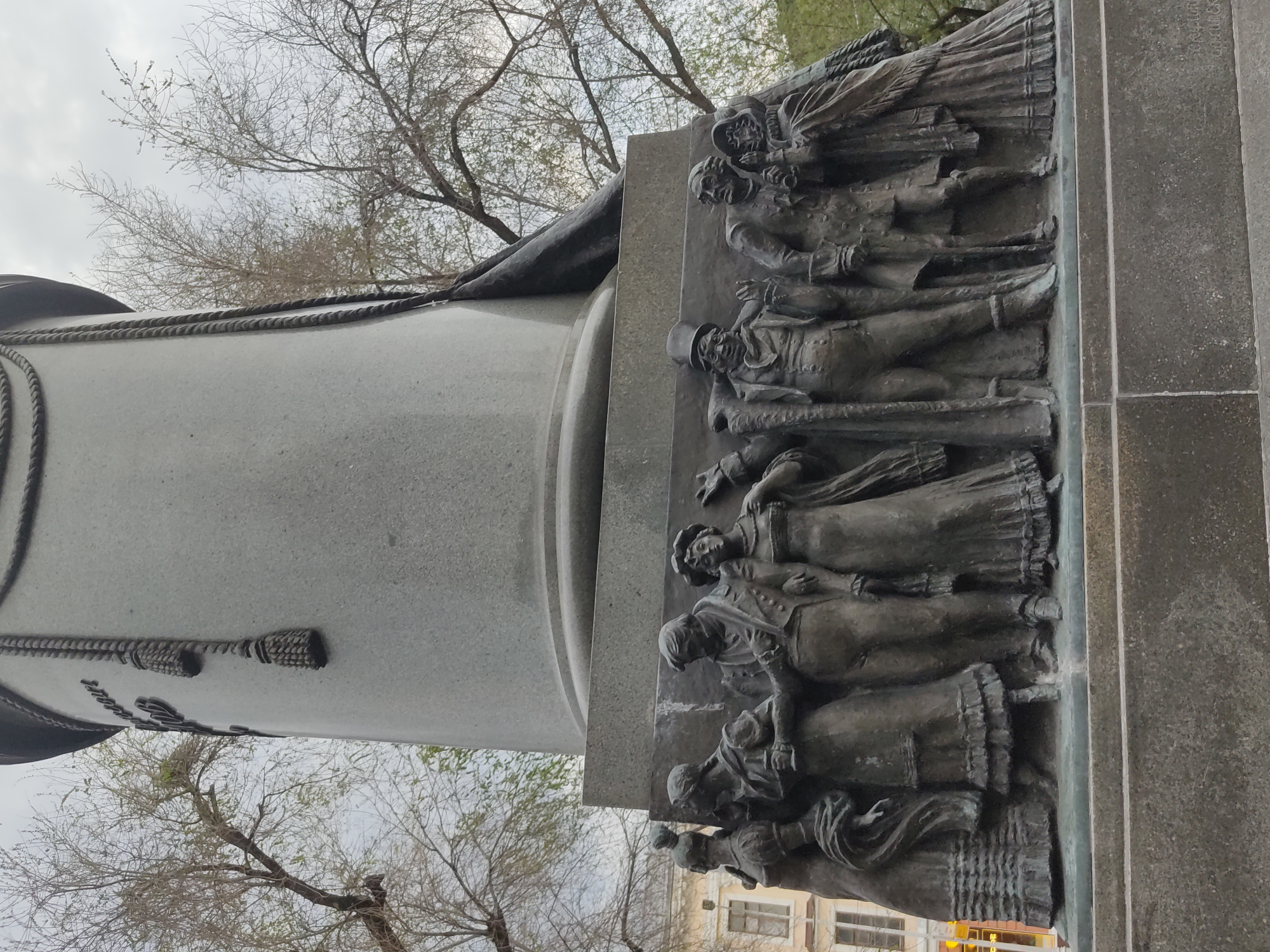 The image depicts a portion of the Monument to Minin and Pozharsky, located on Red Square in Moscow, Russia. The monument's base features a relief sculpture of people in historical dress. The figures appear to be in a procession or gathering. The column of the monument is visible alongside the relief, adorned with a rope motif and tassels. In the background, bare trees and a portion of a building are visible against an overcast sky.