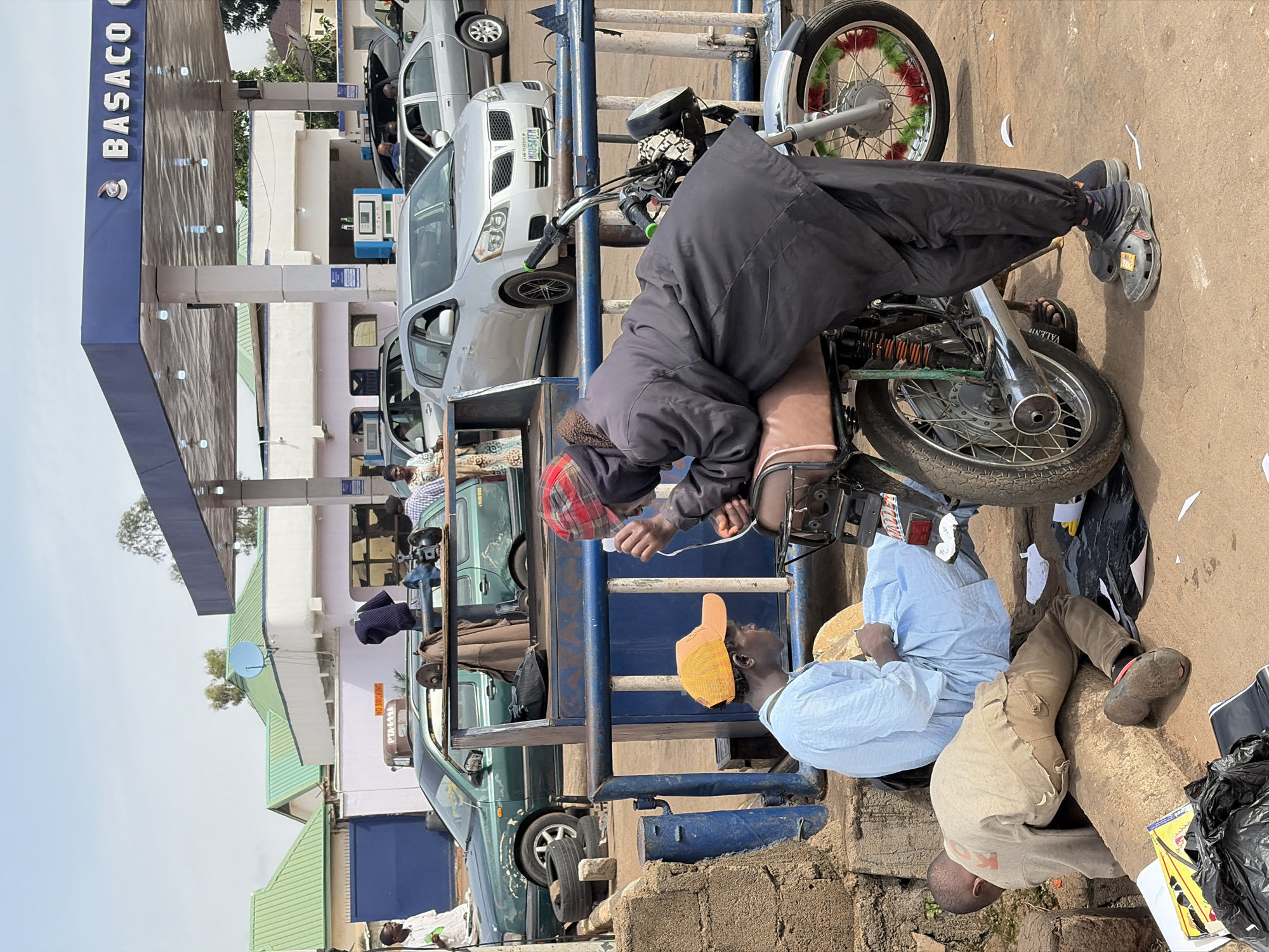 This is a wide shot taken at a BASACO Gas Station in Jos, Nigeria. Three people are visible in the foreground: a person in a blue robe and an orange cap sits on the ground, leaning against a blue barrier; a second person, wearing dark clothing and a red checkered head covering, leans over a motorcycle, seemingly engaged in conversation or an exchange with the person sitting. A third person, wearing a light-colored shirt and dark pants, is partially visible on the ground to the left of the seated individual, appearing to be looking at something on the ground.
In the background, several vehicles are parked, including a green station wagon, a silver sedan, and other cars at the gas pumps. The gas station has a blue and white canopy with lighting. Various objects are scattered on the ground, including papers, plastic bags, and what appear to be tires. The overall environment suggests a busy, outdoor commercial area.