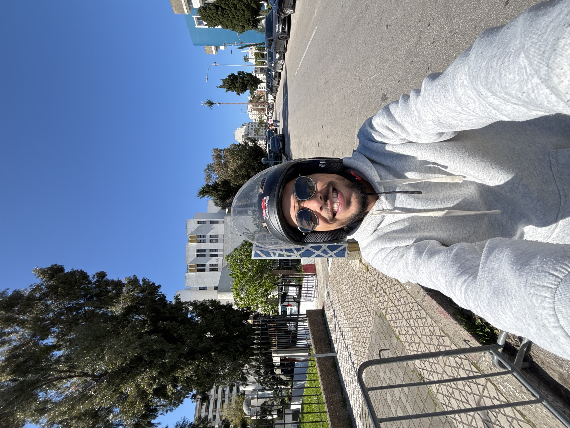A male subject is captured in a selfie-style, eye-level photograph on a clear, sunny day in Tangier, Morocco. The subject, positioned centrally, wears a black motorcycle helmet with a clear visor pushed up, sunglasses, and a light grey hooded sweatshirt with white drawstrings. He has a beard and is smiling, looking directly at the camera. His right arm is extended towards the camera, suggesting he is holding the device.
The scene is set on Avenue Prince Moulay Abdellah in Tangier. To the immediate left of the subject, a grey metal crowd control barrier is visible on a patterned tiled sidewalk, separated by a curb from a narrow strip of grass. Beyond this, a black ornate iron fence encloses a green area, behind which stands a white-domed structure, identifiable as the Mausoleum of Sidi Bou Arraqia. Several white vehicles are parked behind the fence, partially obscured by a large, leafy green tree. Behind this tree and the mausoleum, a multi-story white building with dark rectangular windows and distinctive ochre decorative elements above the windows on its upper floors stands, consistent with the Wilaya de Tanger-Tétouan-Al Hoceïma.
To the right of the subject, an asphalt road with a visible white lane line extends into the distance. Numerous cars, predominantly black and grey sedans, are parked along the right side of the street. In the far distance on the right, two pedestrians are visible walking. Further down the street on the right, a distinctive tall structure resembling a cellular tower disguised as a palm tree is prominent, alongside a street light and a multi-story blue building. The sky above is clear and bright blue.