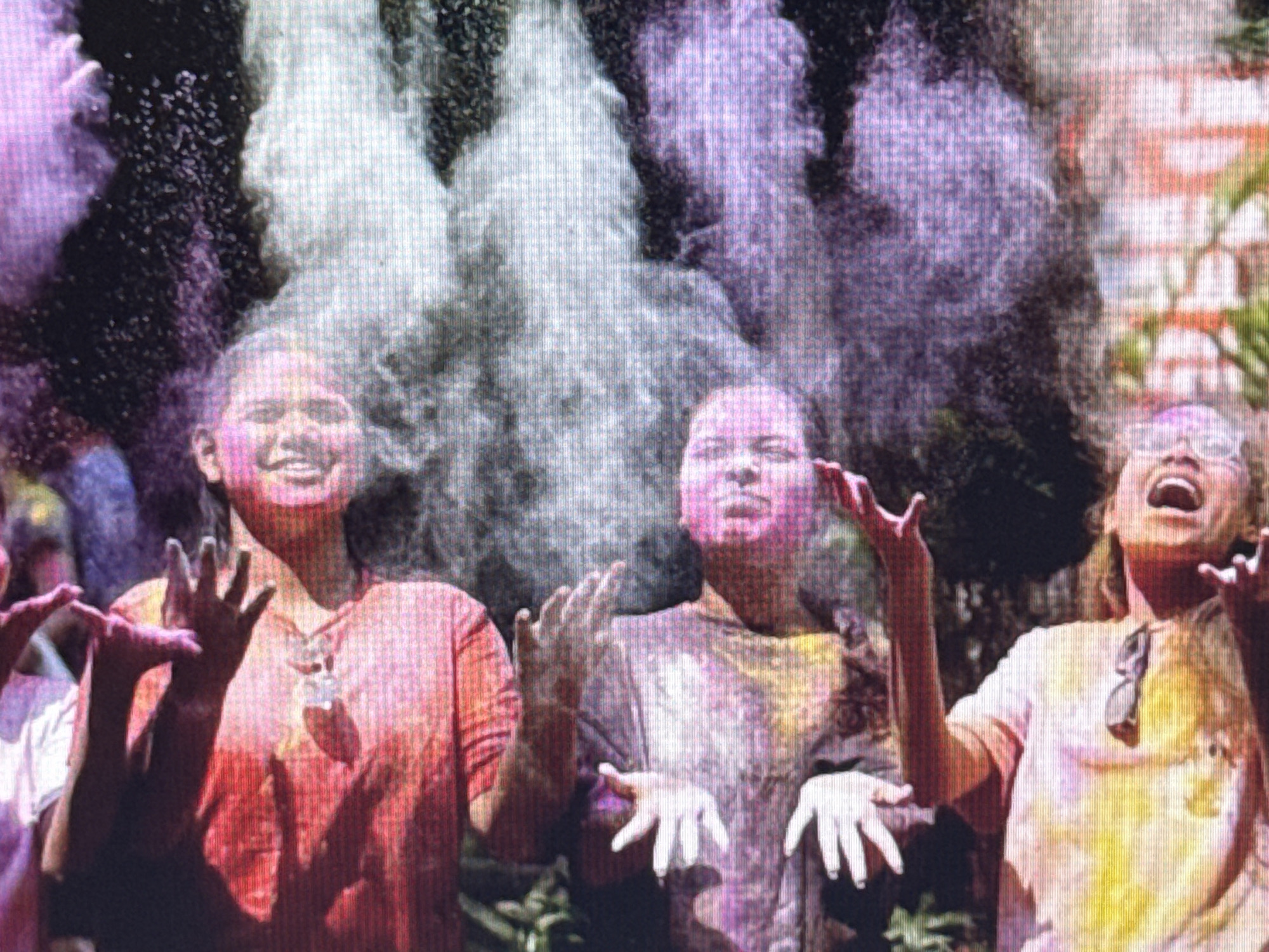 The image depicts a group of individuals celebrating a festival, likely Holi, as they are covered in colorful powders. Three young women are prominently visible. The woman on the left, wearing a red shirt, has her arms raised and hands open, with pink powder on her face and hair. The woman in the center, wearing a purple-tinged shirt, also has her hands outstretched, and her face is partially obscured by pink and white powder. The woman on the right, in a light-colored shirt with yellow and pink powder, is looking upwards with her mouth open, also with powder on her face. Colorful clouds of powder, in shades of pink, purple, and white, fill the air around them. The background appears to be outdoors, with some greenery visible, suggesting a park or open space in a city. The lighting suggests it is daytime.
