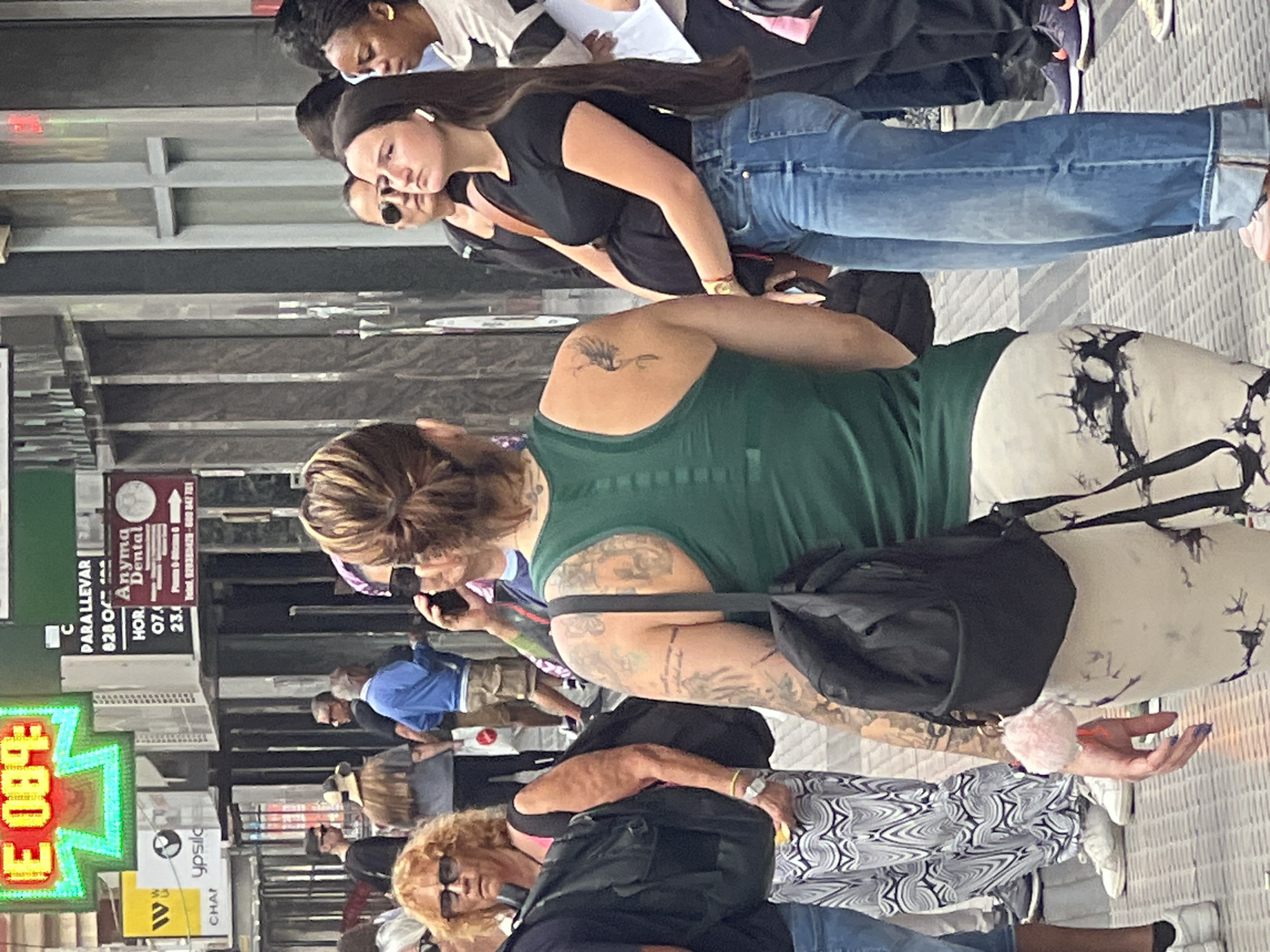 This candid, upright-oriented street photograph captures a bustling commercial area in Las Palmas, Spain, during what appears to be daytime, with a mix of natural and artificial lighting suggesting a covered street or arcade.
In the foreground, a woman with dark blonde hair tied back wears a green tank top, revealing extensive arm tattoos, including a small tattoo of a winged creature on her upper back. She carries a black shoulder bag and wears distinct black and white patterned pants. She is actively looking at her phone, which has a fluffy pink keychain attached to her hand.
Behind her, a young woman with long dark hair, white earbuds, and sunglasses pushed up on her head, wears a black t-shirt and light blue, cuffed jeans. She stands near another person, whose face is obscured by sunglasses. Further back and to the left, another woman with dark skin and braided hair is visible, wearing a white top. In the lower part of the frame, another woman with curly blonde hair and sunglasses on her face wears a black top. Various other people, including a man in a blue shirt, are seen walking or standing in the background.
The setting features concrete overhead structures and various signs, indicating shops or services. On the left, a bright neon sign displays "E 089:". Other visible text on signs includes "C", "PARA LLEVAR", "628 O", "Anyma Dental" with hours "HOR. 07.4 23.4", "ypsilo", "Teiel, 928385425-600 847781", and "W CHAP". The overall mood is neutral, depicting people engaged in their daily routines on a city street.