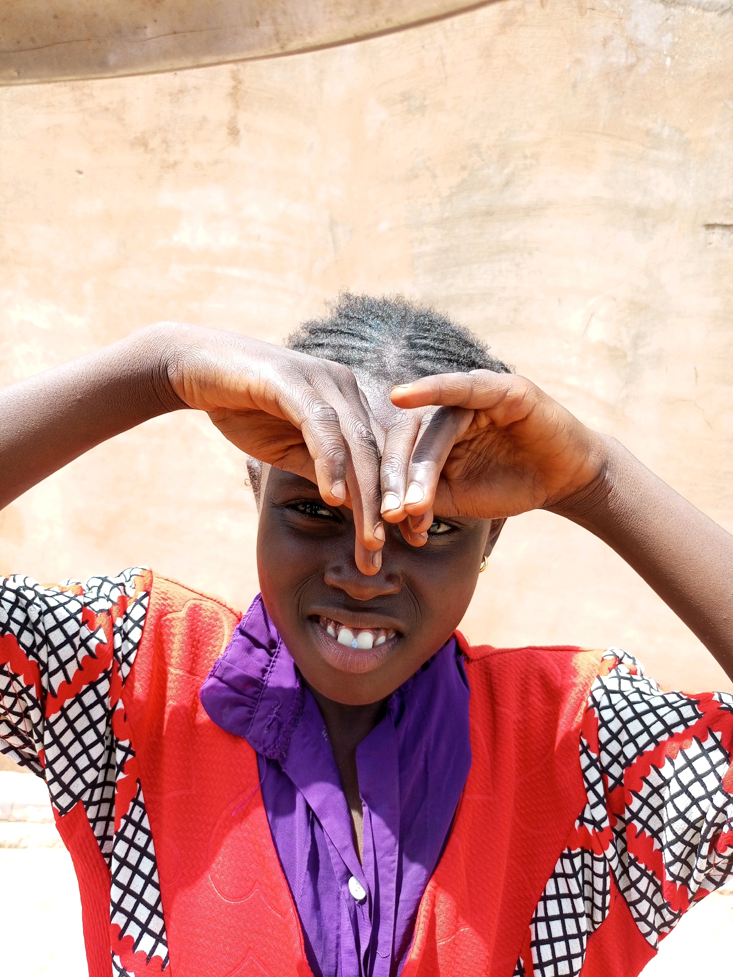 In a medium shot, a young African child, with dark skin and braided hair, playfully covers their eyes with their hands. The child wears a vibrant red shirt with a black and white geometric pattern on the sleeves and purple collar, over a purple button-up shirt. They are smiling, revealing their teeth. The background is a textured, light brown wall. The scene is captured outdoors under bright light, suggesting it is daytime. The location is identified as Tempure, Nigeria.