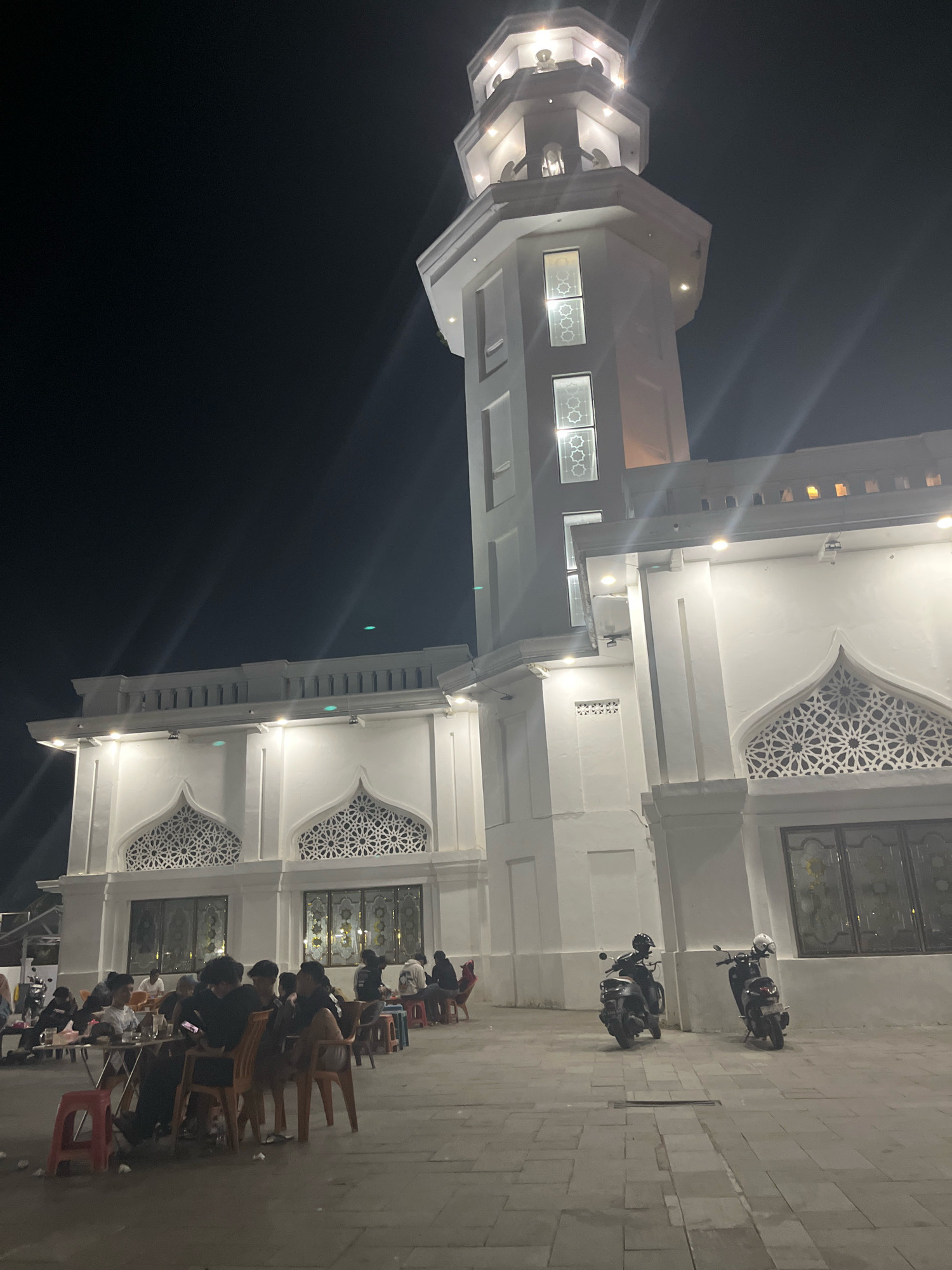 The image depicts the exterior of a white mosque at night in Sengkang, Indonesia. A prominent minaret, illuminated by exterior lights, rises to the left of the frame, featuring decorative windows with intricate geometric patterns. The main mosque structure also exhibits similar decorative window designs above large glass panes. In the foreground, several people are seated at tables and chairs outside the mosque, appearing to be socializing or dining. Two motorcycles are parked on the tiled ground in front of the mosque. The night sky is dark, with some lens flares present from the artificial lighting.