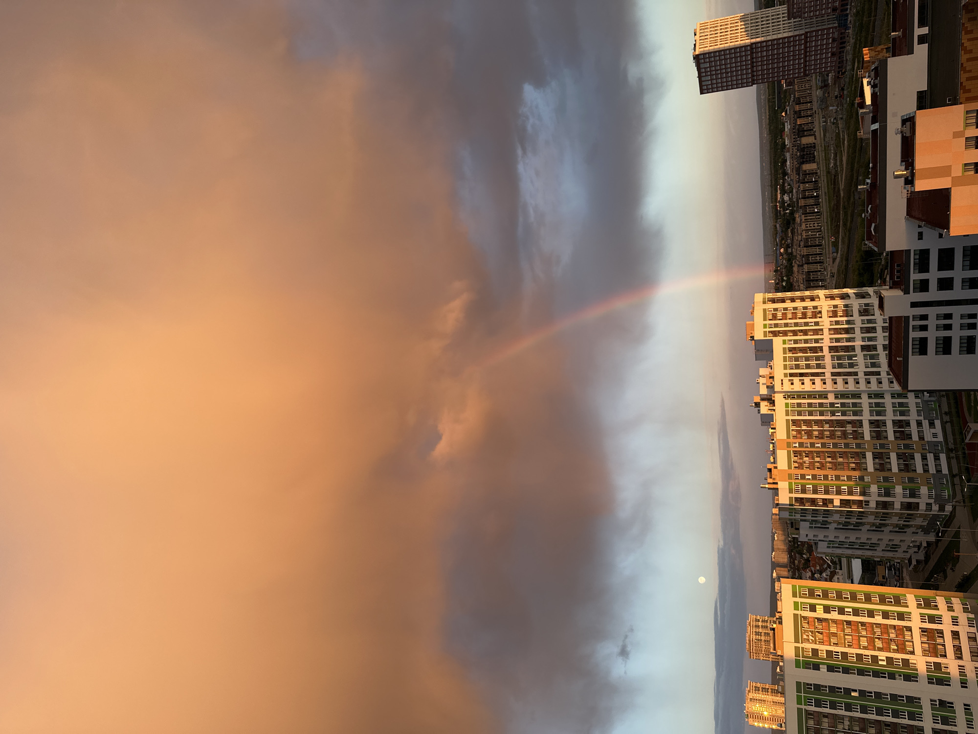 The image captures a panoramic view of a cityscape, likely Yekaterinburg, Russia, judging by the architecture. Several high-rise apartment buildings are visible, displaying a variety of colors and architectural styles. A prominent rainbow arcs across the sky, intersecting with the cityscape. The sky itself is divided into two distinct sections: one is filled with orange-tinged clouds, suggesting sunset or sunrise, while the other is dominated by darker, stormier clouds. In the lighter part of the sky, a bright, circular object resembling the moon is visible. The photograph appears to have been taken from an elevated position, offering an expansive view of the surrounding area. The buildings are reflecting the light of the sun, suggesting the time of day is either early morning or late afternoon. The image is oriented vertically.