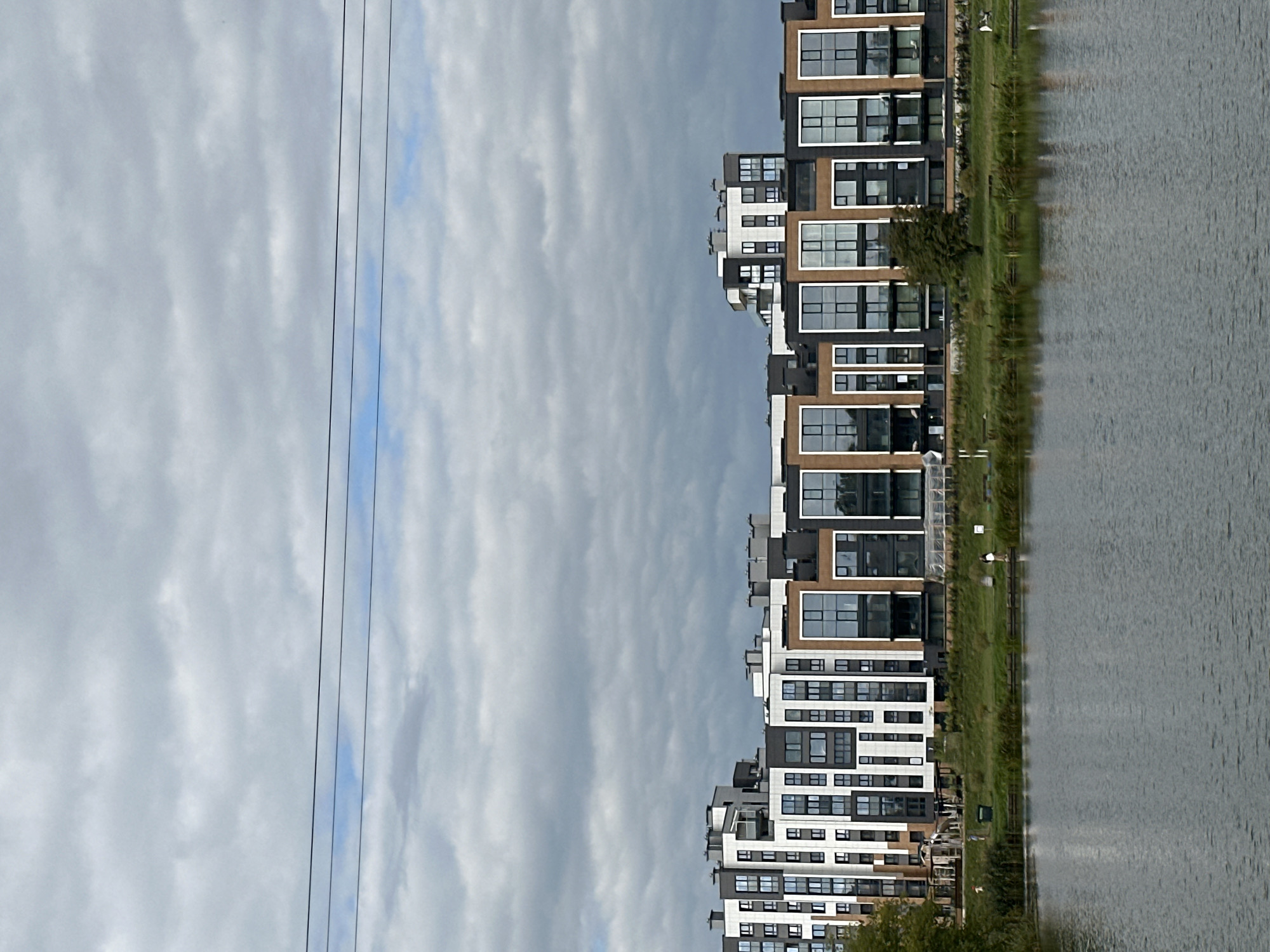 The image depicts a waterfront view of modern apartment buildings. The buildings are multi-story structures with a contemporary architectural design, featuring a combination of white, dark gray, and brown facades, with large windows. They are situated along a grassy embankment that slopes down to a calm body of water, likely a lake or river. Several trees are visible on the embankment, and a few small figures of people can be seen walking or standing near the water's edge. The sky is overcast with heavy gray clouds. Three thin, dark power lines are stretched horizontally across the upper portion of the image.