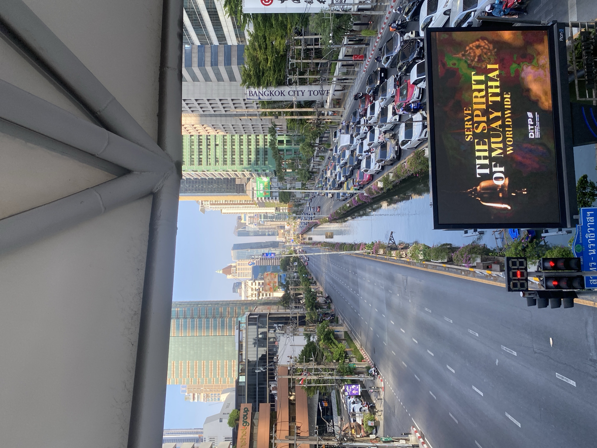 This image, taken from an elevated walkway, captures a wide-angle urban street scene in Bangkok, Thailand, under clear daytime skies. The perspective looks down a long road flanked by city infrastructure.
On the left side, a long, narrow canal runs parallel to the road. Adjacent to the canal, a section of the road is heavily congested with numerous cars parked or moving slowly, suggesting a traffic jam. Behind this, several modern high-rise buildings dominate the skyline, including one clearly marked "BANGKOK CITY TOWER" and another displaying "photgroup". Green trees and foliage are visible along the canal and between buildings.
The center of the image features a multi-lane road that appears largely clear of traffic for a considerable distance, stretching into the background where more city buildings are visible under a bright sky.
On the right side of the frame, a large digital billboard is prominent, displaying the text: "SERVE THE SPIRIT OF MUAY THAI WORLDWIDE." It also includes "DITP" and "Plan-B" branding. Below the billboard, a traffic light shows red signals, accompanied by a digital countdown timer displaying "888". Adjacent to the traffic light, Thai text is visible, which translates to "Intersection Sathorn-Naradhiwas," indicating the specific location within Bangkok. The foreground reveals the gray metallic structure of the overhead pedestrian bridge from which the photograph was captured.