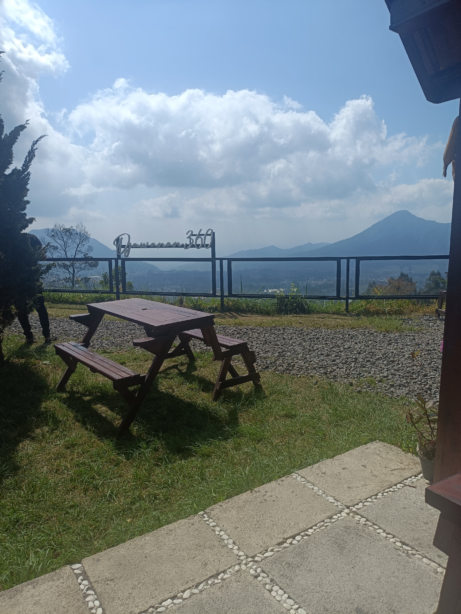 The image shows an outdoor seating area with a wooden picnic table and benches on a grassy lawn. In the background, there is a panoramic view of mountains and valleys under a partly cloudy sky. A metal fence with glass panels separates the seating area from the view. A sign with the text "Pemandangan 360" is visible above the fence. There is also a person standing near a tree on the left side of the image. The foreground features a concrete pathway decorated with white pebbles. The scene appears to be a scenic viewpoint or a cafe overlooking a natural landscape in Kopeng, Indonesia.