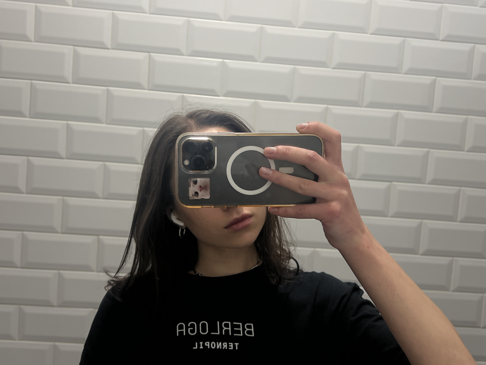 A young person with dark hair is taking a mirror selfie. They are wearing a black t-shirt with white text on it. They also have white earbuds in their ears and a phone in their right hand, which is obscuring their face. The background appears to be a white tiled wall. The text on the t-shirt is reversed in the reflection, and appears to read "TERNOPILL" on the bottom line and "BERLOOC" on the top line. This indicates the location is Ternopil, Ukraine.