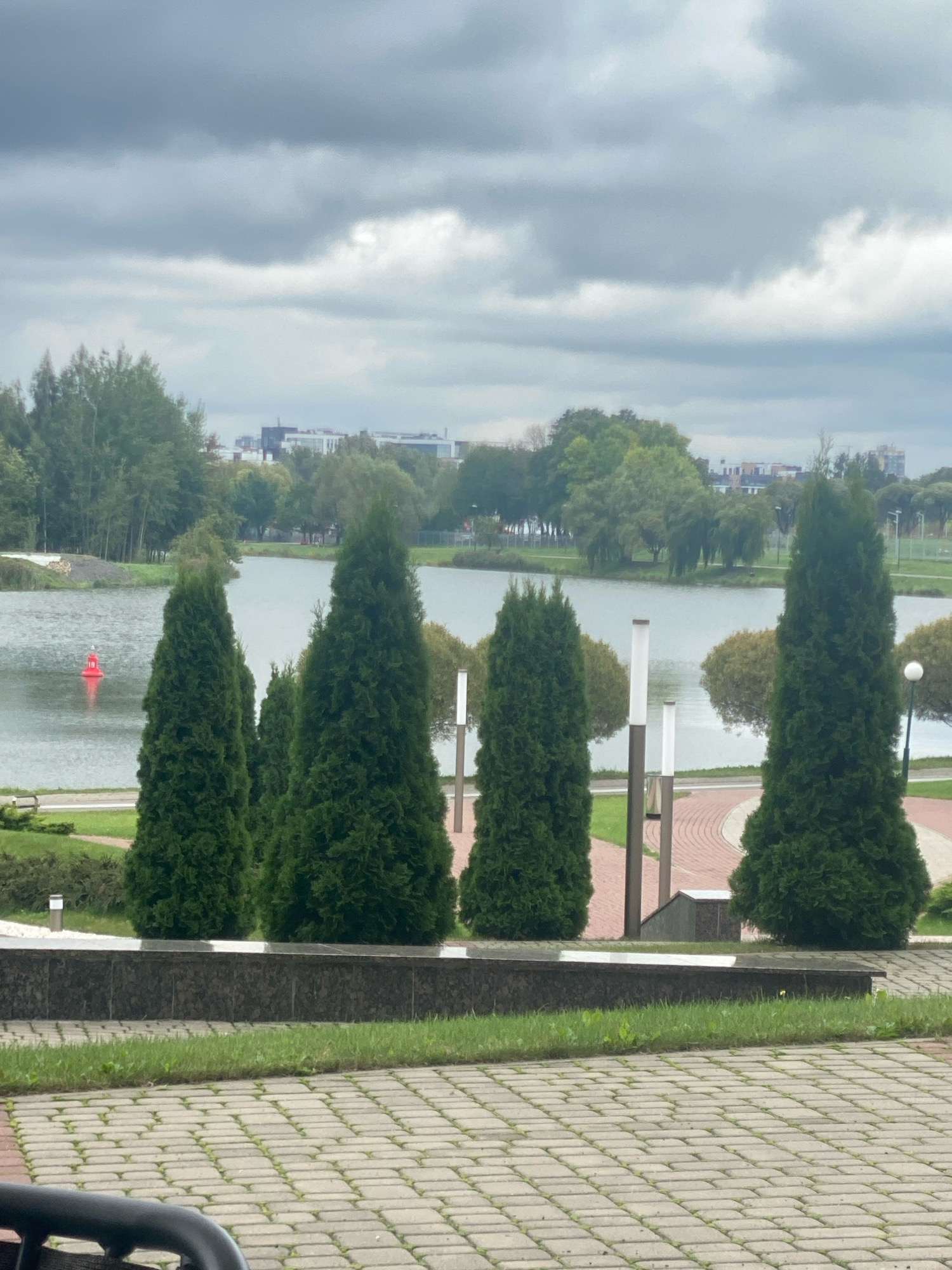 The image shows a cloudy day in a park in Minsk, Belarus. In the foreground, a paved walkway made of interlocking rectangular stones leads to a stone retaining wall. Beyond the wall, grass and several tall, conical evergreen trees are visible. A body of water, likely a lake or river, is in the middle ground, with a red buoy floating in it. Across the water, trees and buildings can be seen, suggesting an urban park setting. The sky is overcast with heavy grey clouds. There are no people or animals visible in the image. The overall atmosphere is subdued due to the weather conditions.