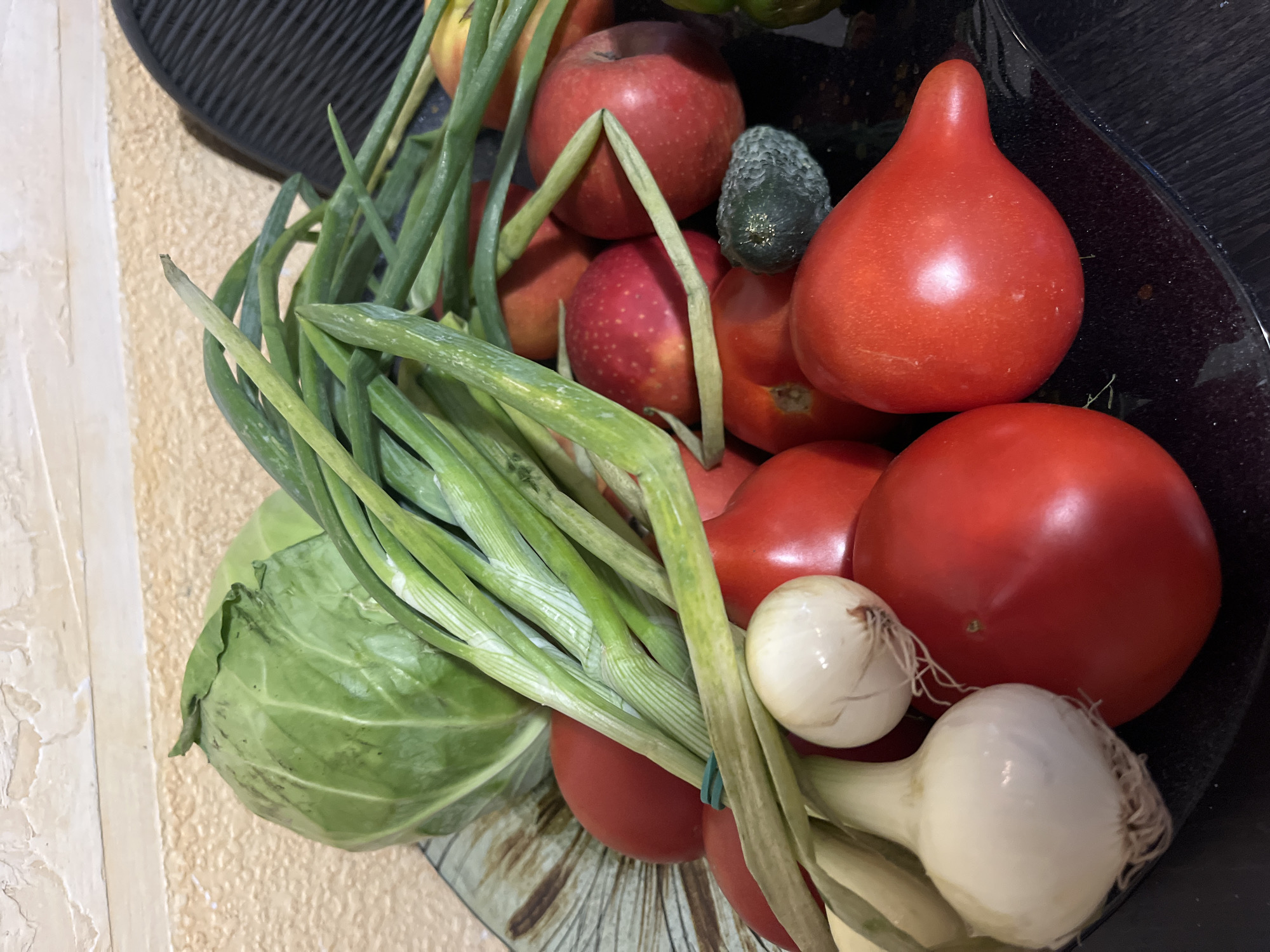 A collection of fresh produce is displayed in a bowl on a countertop, likely in a domestic kitchen in Tarnivtsi, Ukraine. The bowl, made of a ceramic material with a pattern of brown and grey streaks, contains a variety of vegetables and fruits. Prominently featured are several ripe red tomatoes, some with a classic round shape and others with a more elongated, pear-like form. Among the tomatoes, a head of green cabbage is visible, its leaves tightly packed. A bunch of green onions, tied together with a green rubber band, lies across the other produce. Also present are two small white onions with visible roots. In the background, there are a few red apples with subtle yellow and green undertones, and a small, bumpy, dark green object that appears to be a cucumber. The countertop itself is a dark, reflective surface, possibly granite or a similar material. The background wall has a textured finish, a mix of light beige and cream tones. The scene suggests a recent harvest or grocery shopping, with the fresh ingredients ready for preparation.