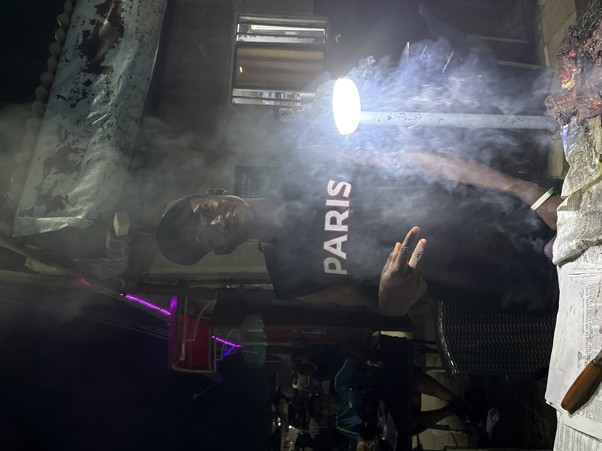 A man wearing a black sleeveless shirt with "PARIS" in white lettering and a black baseball cap is standing outdoors at night. He is looking towards the camera and making a peace sign with his right hand. Smoke is billowing around him, illuminated by a bright light source. In the foreground, a grill with meat is visible, placed on newspapers. In the background, there are blurry figures of people seated at tables and buildings with visible signs and lights, suggesting a marketplace or roadside food stall in Abuja, Nigeria. The atmosphere is smoky and dimly lit, typical of an evening street food setting.