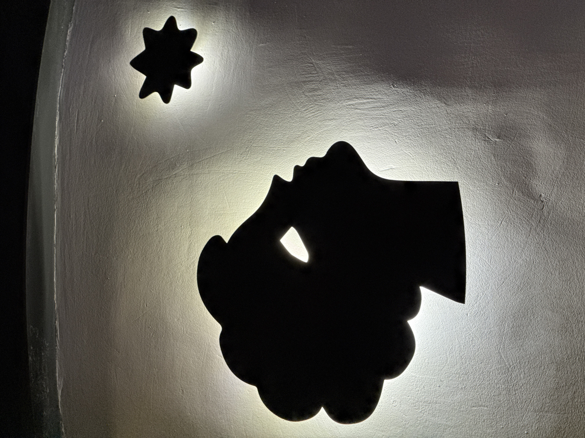 This media file presents a backlit decorative art piece on a textured, light-colored wall. Two prominent black silhouettes dominate the frame: an eight-pointed star positioned in the upper left, and a larger, more complex shape below it.
The larger silhouette distinctly features the profile of a human face, with a visible nose, mouth, and chin, looking upwards and to the left, seemingly gazing towards the star. This facial profile merges into a broader, rounded, almost cloud-like form, which contains a small, irregular cutout glowing brightly within its mass.
The strong backlighting creates a luminous halo effect around the edges of both silhouettes, highlighting their shapes and casting a warm glow on the uneven, possibly plastered or stuccoed wall surface. The light is brightest immediately around the cutouts, gradually diminishing across the wall, with a darker, vertical strip visible along the far left edge of the image.
The scene is static, representing a decorative installation rather than an active event. The composition, with the face directed towards the star, suggests themes of contemplation, aspiration, or wonder. The absence of natural light implies an indoor setting where artificial lighting creates this visual effect. There is no visible text in the image. The location context for this piece is Yerevan, Armenia.