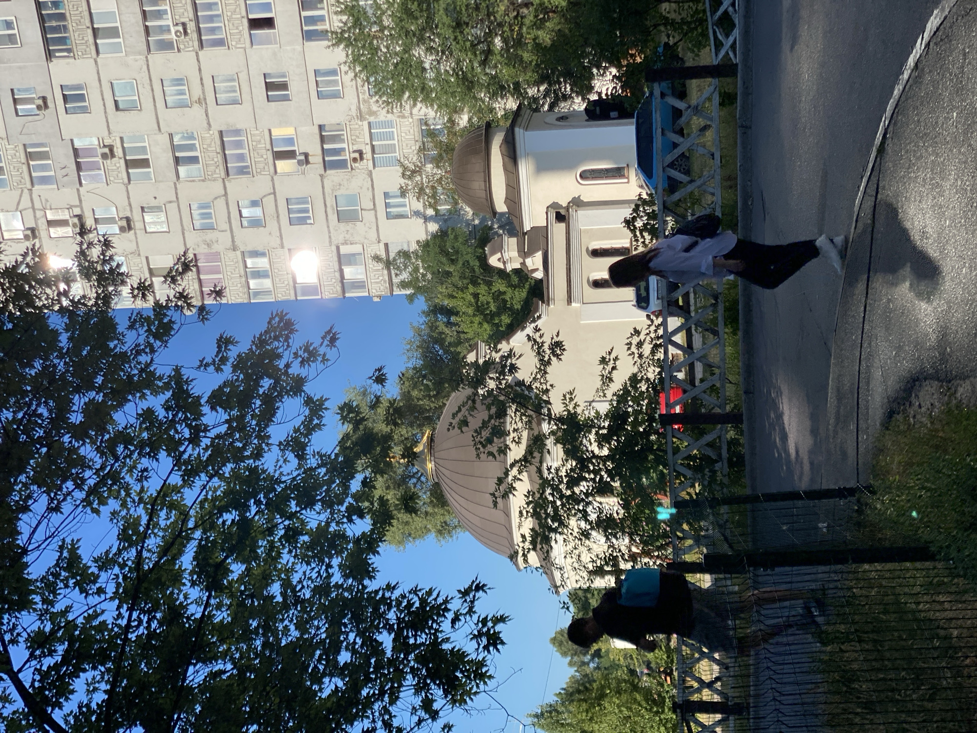 In a city scene of Unknown, Unknown, an outdoor view features a multi-story apartment building and a church-like structure with a dome. Sunlight reflects brightly off the apartment building's windows. Green trees and foliage are present throughout the scene, with large tree branches and leaves in the foreground obscuring parts of the view. In the middle ground, a man wearing a dark shirt and shorts, carrying a light blue bag on his back, walks with his head down. To his right, a woman in a light-colored shirt and dark pants walks away from the viewer, carrying a dark backpack. A white fence with a crisscross pattern runs across the middle ground. Several cars are visible behind the fence. The ground appears to be asphalt.