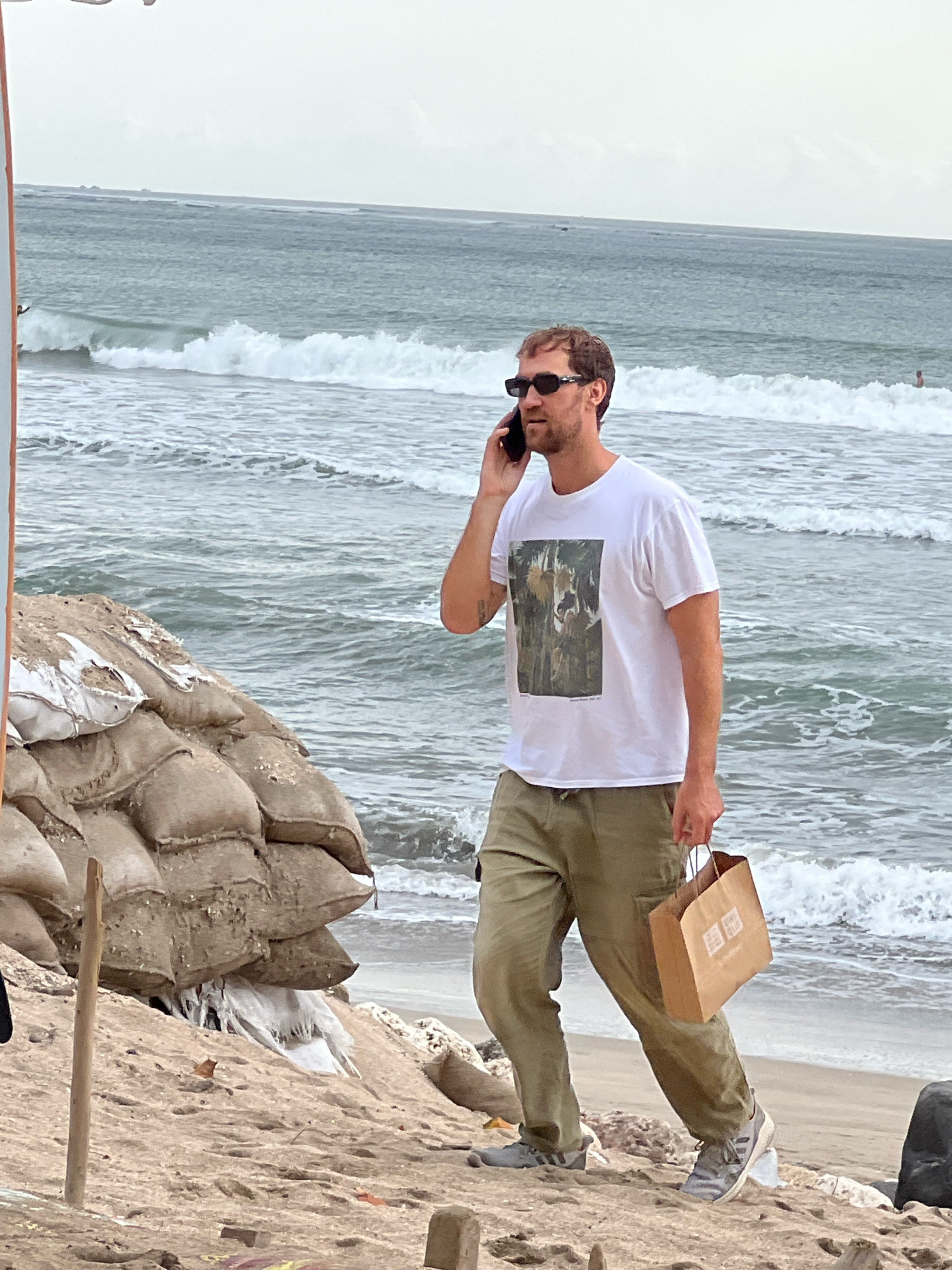 A Caucasian male wearing sunglasses, a white t-shirt with a graphic print, and green cargo pants is walking on a beach in Kuta, Indonesia. He is holding a paper shopping bag and talking on a mobile phone. The background shows the ocean with waves and a cloudy sky. Sandbags are piled up on the left side of the frame, suggesting possible coastal erosion control or construction. The ground is sandy and appears to be a public beach.