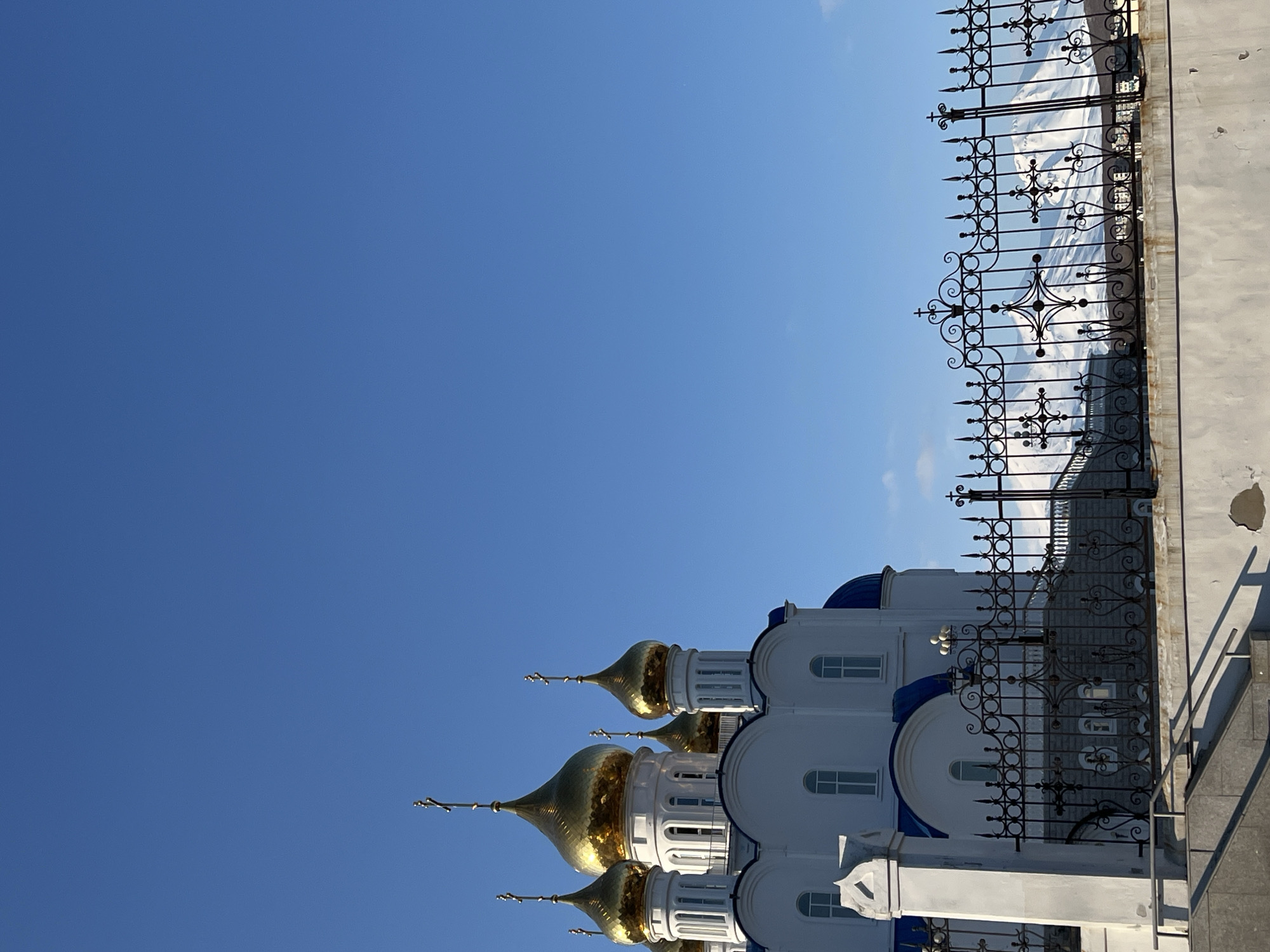 The image shows a low-angle view of the Holy Trinity Cathedral in Petropavlovsk-Kamchatsky, Russia. The cathedral features white walls with blue accents, golden domes, and crosses. A black wrought-iron fence runs along the right side of the image, partially obscuring the background, which appears to be a snowy landscape. The sky is a clear, vibrant blue.