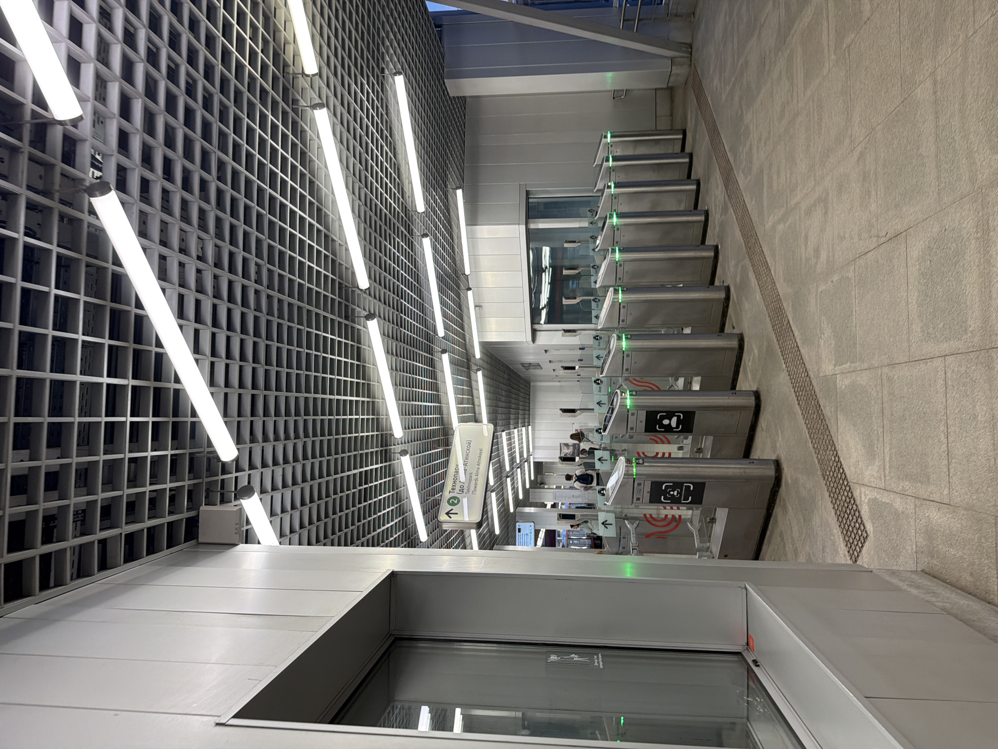 An indoor, brightly lit perspective of a metro station entrance, facing a line of modern, silver turnstiles. The turnstiles have green indicator lights, suggesting they are operational and ready for use. Each turnstile features an icon resembling a person or a face, likely indicating the method of access. Ahead of the turnstiles, a directional sign points to "Технопарк (Tekhnopark) (towards Alma-Atinskaya)". In the background, a few individuals are visible, seemingly in the process of passing through or waiting to pass through the turnstiles. The ceiling of the station is a grid of metal, with long, fluorescent lights running parallel to each other. The walls are a smooth, metallic silver. A large window is visible on the left side of the frame, offering a glimpse into an adjacent space. The floor is tiled with large, light-colored square tiles, and a tactile paving strip runs along the front of the turnstiles, indicating accessibility features.