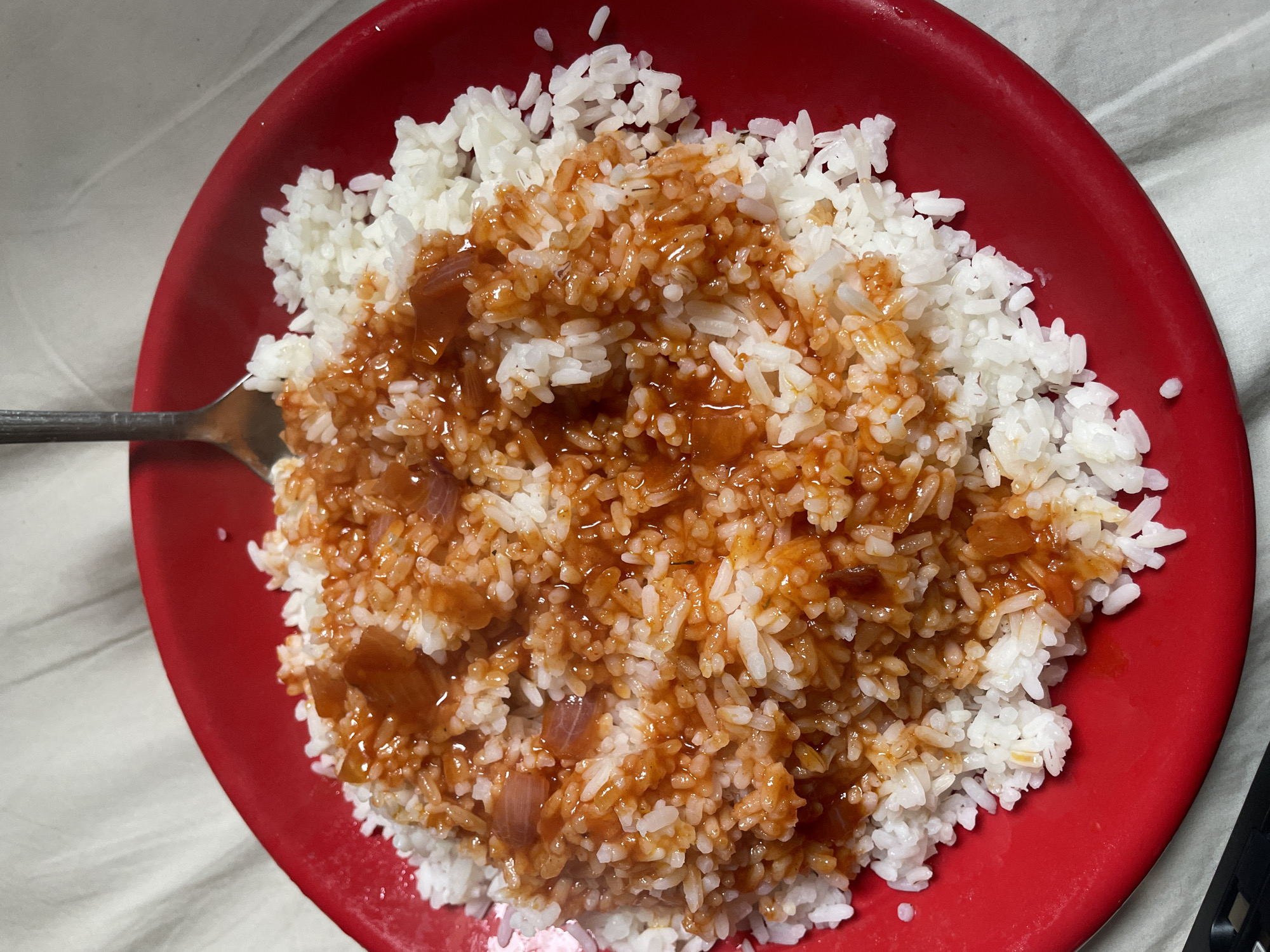 A red plate filled with white rice and a reddish sauce with visible pieces of onion sits on a white, textured surface. A fork is partially submerged in the rice, with its tines digging into the dish. The lighting suggests an indoor setting. The image does not provide any information about the location in Safari, Nigeria.