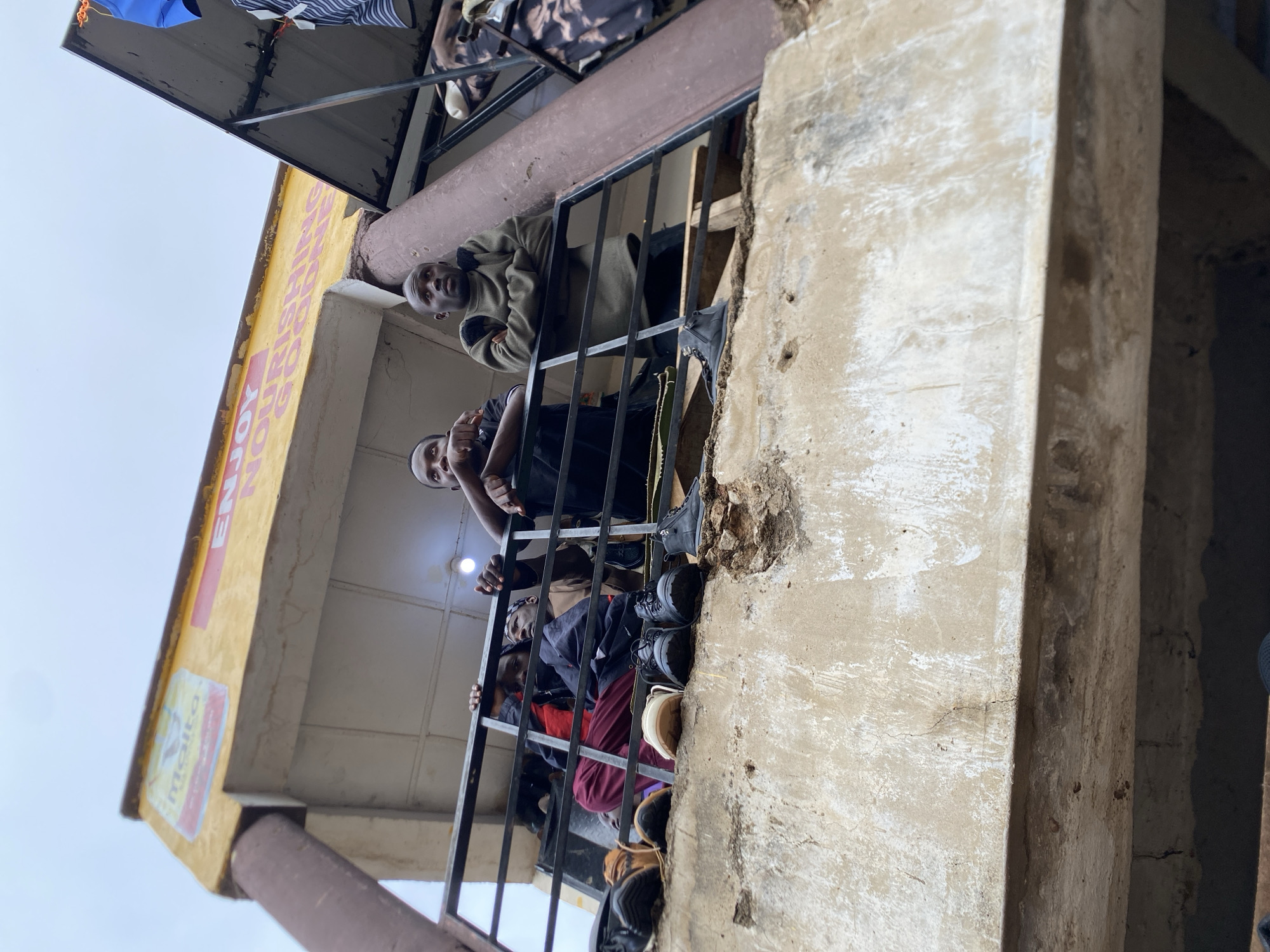 The image shows a group of people on a balcony in Bukuru, Nigeria. The balcony has a black metal railing and is attached to a concrete building. The building has a yellow overhang with the words "Enjoy Nursing Home" visible. The people on the balcony are looking down at something. The photograph appears to be taken from a low angle, looking up at the building.