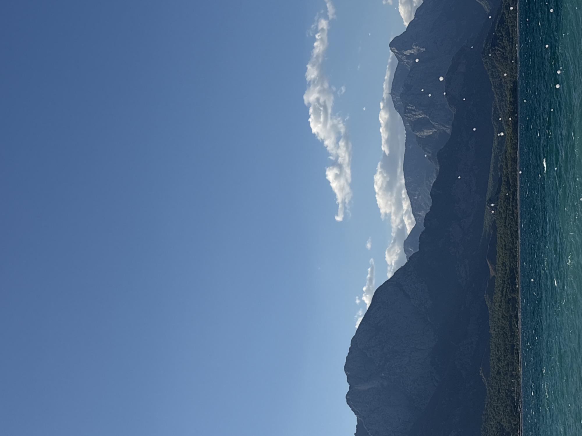 A wide-angle, vertical shot captures a serene landscape in Kemer, Turkey. The upper half of the frame is dominated by a vast, clear blue sky, interrupted by scattered white clouds. Below the sky, a range of dark, rugged mountains forms the horizon. The mountains are densely covered with lush green trees in the foothills. In the foreground, the turquoise waters of the sea are visible, with gentle waves creating a textured surface. Small, white, circular artifacts are scattered throughout the image, particularly over the mountains and sea. The overall scene evokes a peaceful, natural environment.