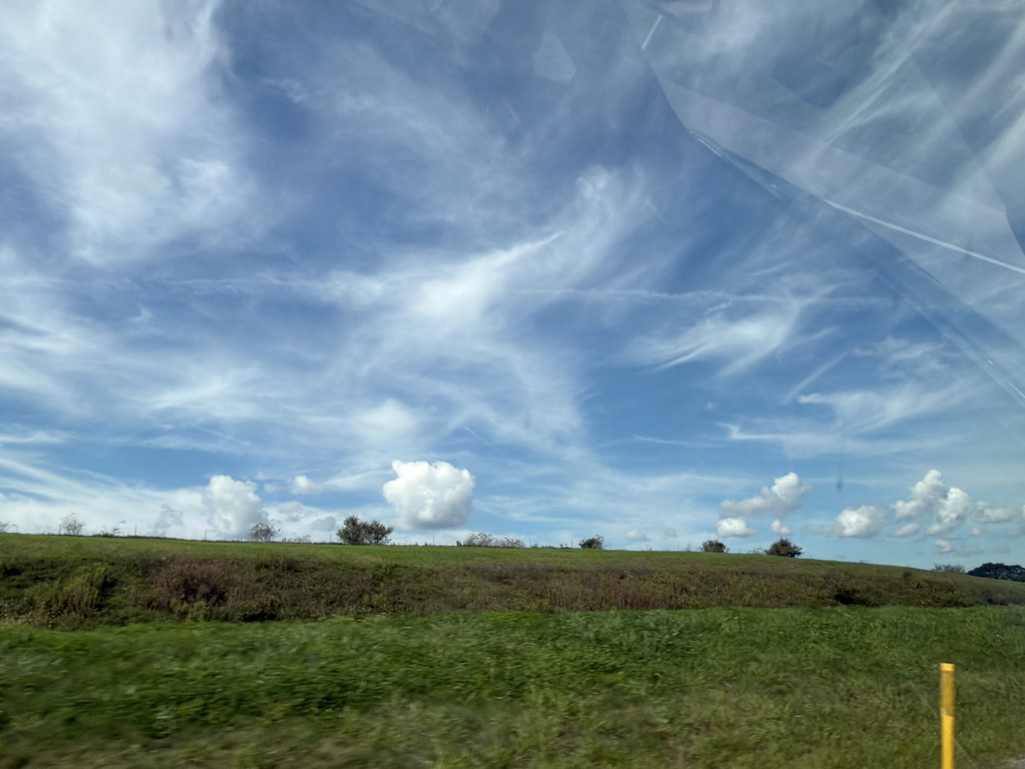 The image depicts a wide, grassy field under a vast sky with wispy white clouds. The field slopes upwards, forming a gentle hill in the background. Scattered trees and bushes dot the crest of the hill. The foreground is dominated by lush green grass, which appears slightly blurred, suggesting movement. A bright yellow post stands on the right side of the frame, partially visible in the foreground. The sky is a vibrant blue, with intricate patterns of cirrus clouds stretching across it. Reflections on a glass surface, likely a window, are visible in the upper portion of the image, obscuring parts of the sky. The scene appears to be an open, natural landscape, possibly a park or rural area. No people or buildings are present in the frame.