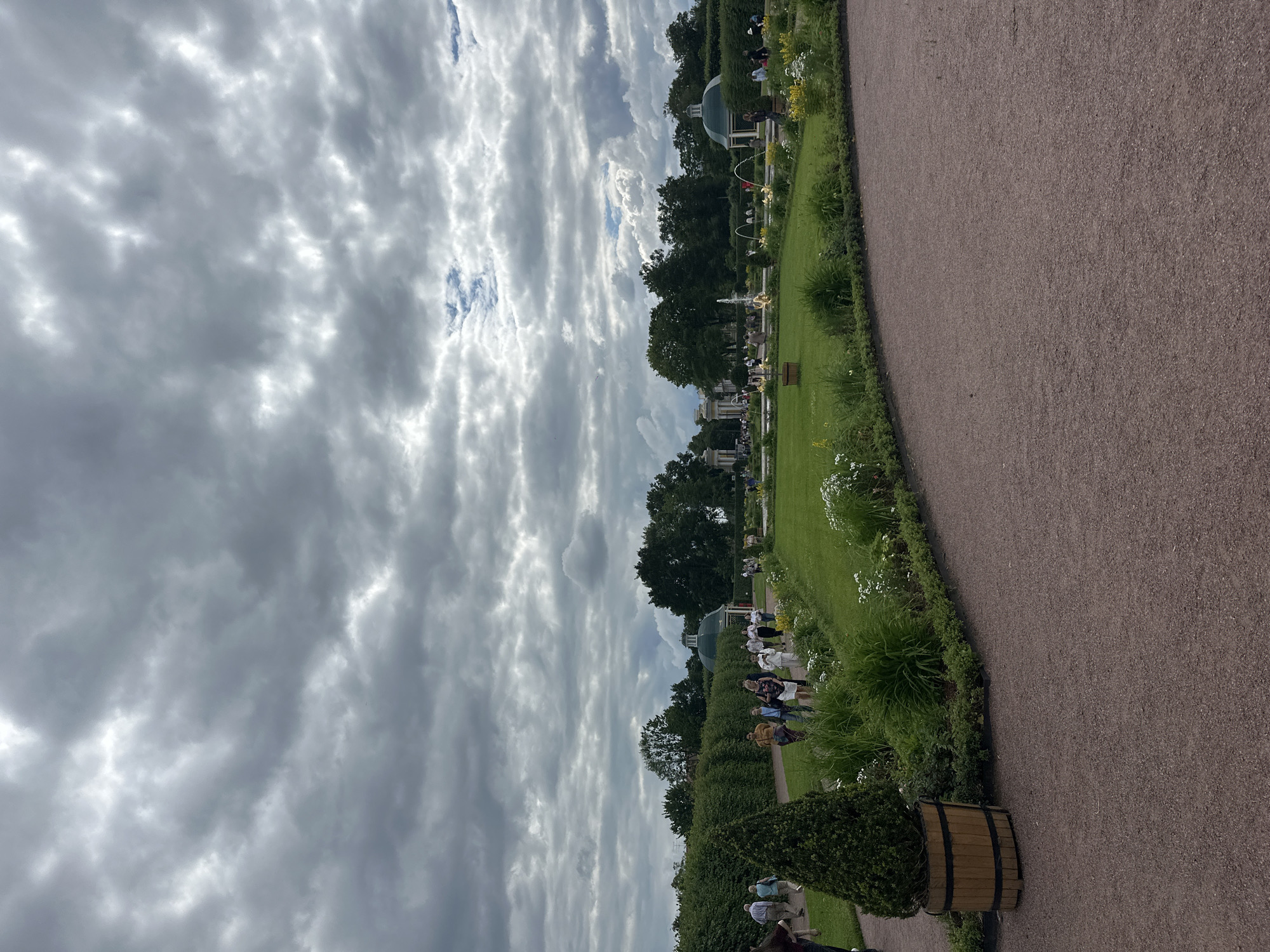 This is a photograph of a garden pathway in Peterhof, Russia, under a cloudy sky. The pathway, made of reddish-brown gravel, curves from the lower right to the upper left of the frame. To the left of the pathway is a large, manicured evergreen shrub in a wooden planter. Beyond this shrub, a neatly trimmed green hedge forms a boundary for a lawn area.
On the lawn, several people are walking along a paved path, appearing to be tourists. In the middle ground, there are fountains with water jets spraying into the air, and a domed green pavilion is visible. More trees and some white and yellow flowers are scattered throughout the garden. In the far background, there are larger buildings and more trees. The overall scene depicts a public garden with visitors enjoying the landscape.
