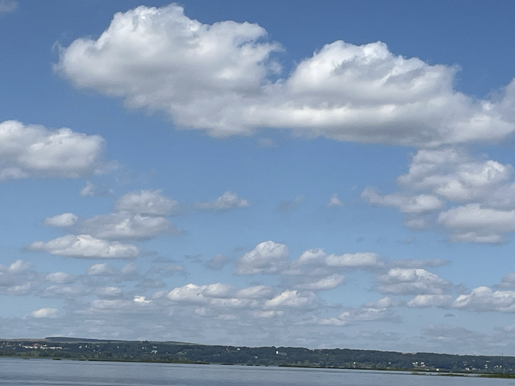 A wide shot captures a serene view of the sky over Shumeika, Russia. The sky is a vibrant blue, filled with a scattering of fluffy white cumulus clouds. Below, a calm, expansive body of water stretches across the lower portion of the frame, reflecting the soft light. In the distance, a lush green landscape with a dense treeline borders the water, punctuated by hints of distant buildings, suggesting a small settlement nestled along the shore. The overall scene conveys a peaceful and natural atmosphere.