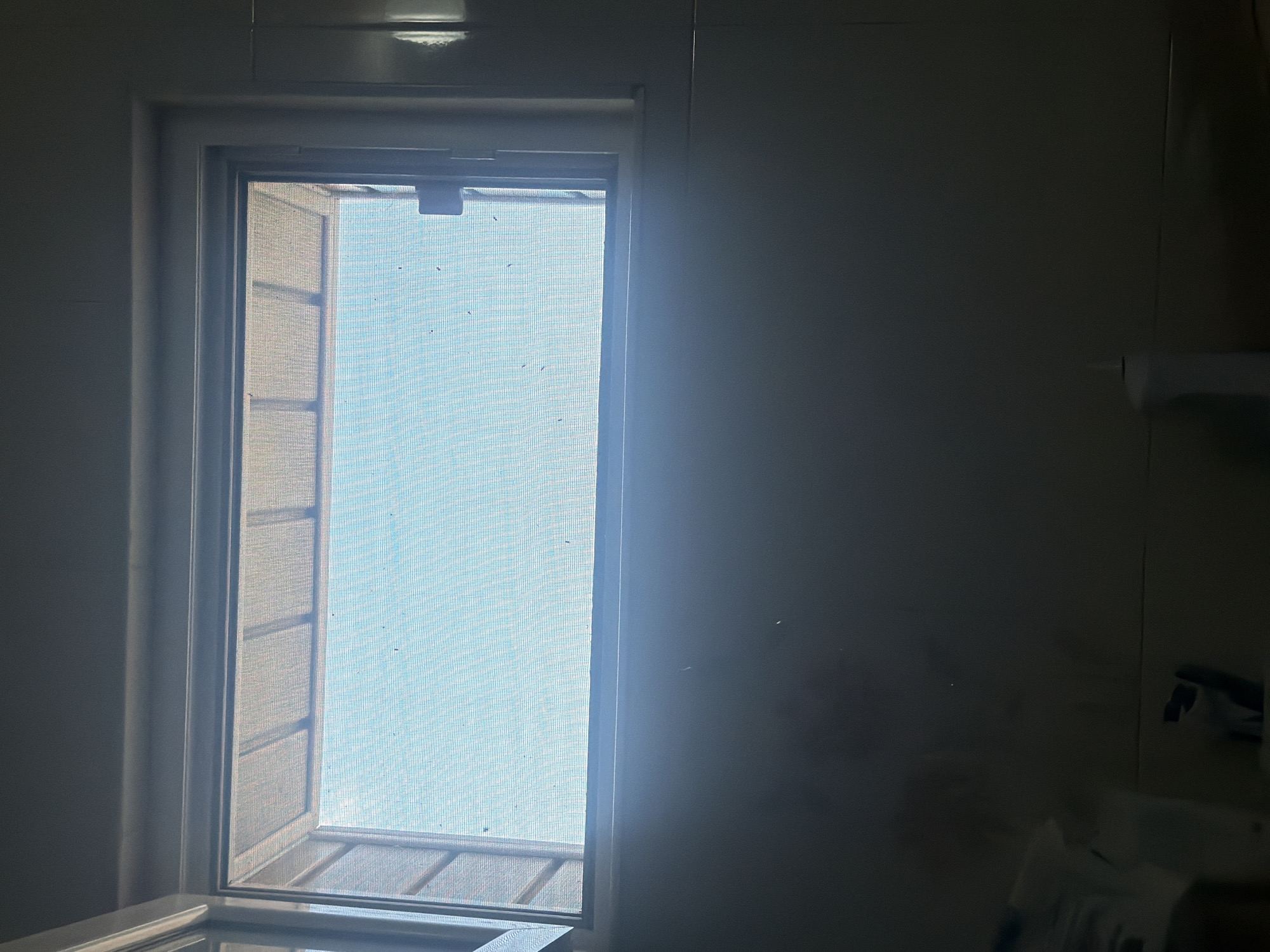 The image displays an interior scene, likely in a room in Iskar, Bulgaria, focusing on a tall, rectangular window. Bright daylight streams through the window, backlighting the interior and creating a strong contrast between the luminous exterior view and the dimly lit room. The window is covered with a fine mesh screen, revealing a bright, clear, blueish sky, indicating daytime and good weather conditions. Small specks of dust or debris are visible on the screen. A dark, rectangular fixture, possibly a latch or part of a ventilation system, is located at the top center of the window frame.
The wall to the left of the window is plain and light-colored. The right side of the room is heavily shadowed; however, faint details are discernible. A horizontal fixture, possibly a towel bar or shelf, with a pale object hanging from it, is visible on the wall in the upper right quadrant. In the lower right corner, indistinct, very dark shapes suggest household items on a surface. The scene is static, devoid of people or specific activities, and no text is visible. The overall impression is one of quiet functional space.