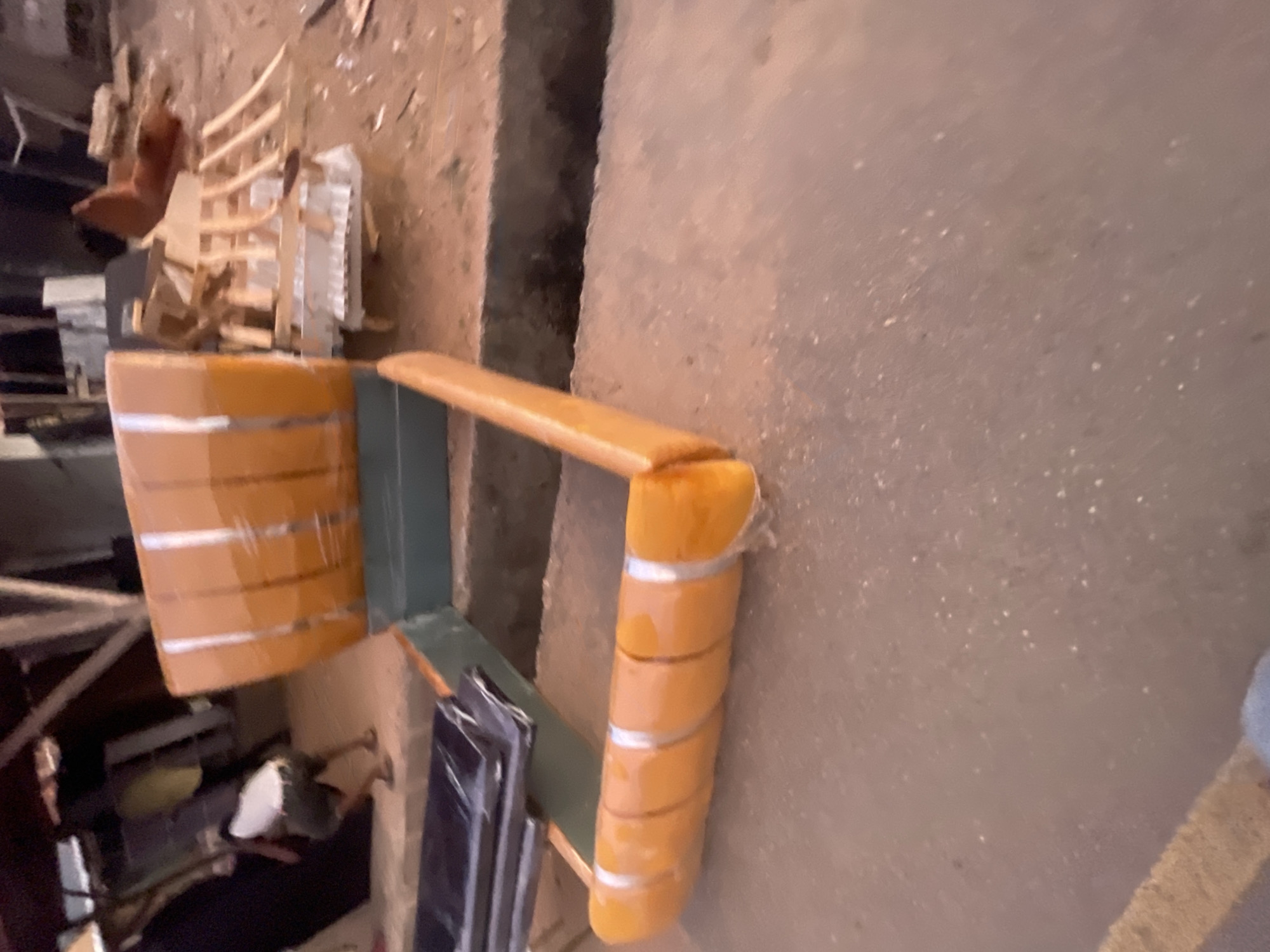 The image depicts what appears to be a workshop or storage area in Lokoja, Nigeria. An orange-colored object with horizontal lines, possibly furniture or a component of a piece of furniture, is prominently displayed. Wooden structures and what looks like disassembled furniture parts are visible in the background. The environment suggests an industrial or artisanal setting. No people are discernible in the shot.