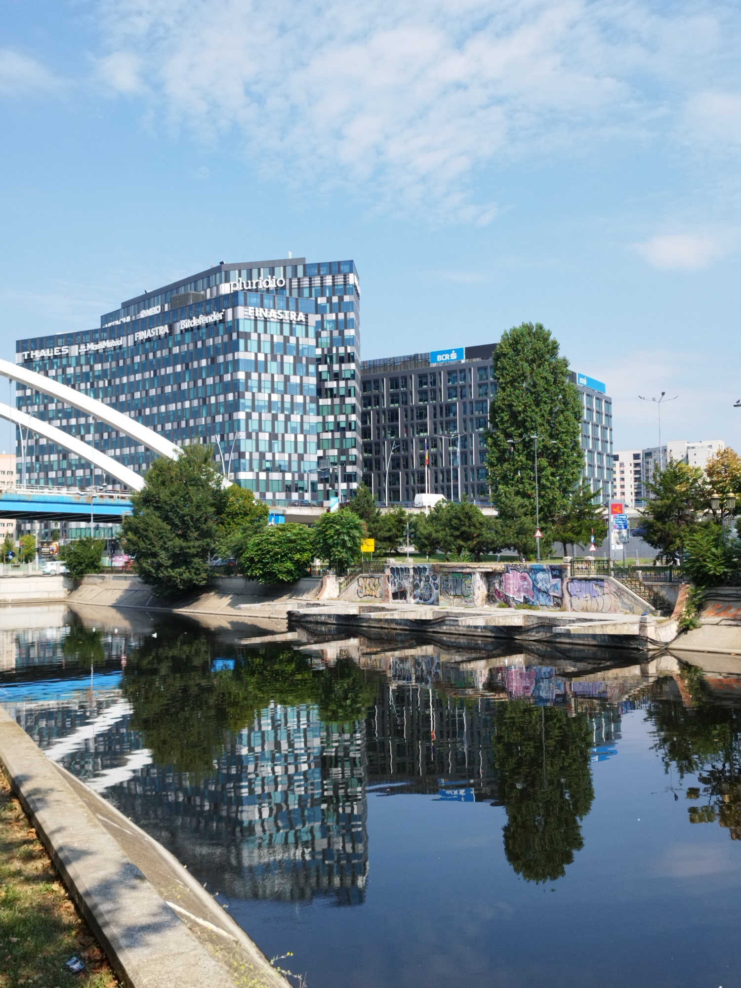 A vertical, medium-shot photograph captures a cityscape in Bucharest, Romania, dominated by a large, modern office building with a patterned facade. The building, with a stepped architectural design, features a glass exterior reflecting the blue sky and white clouds. Visible text on the building includes "pluridio," "THALES," "MassMutual," "FINASTRA," and "Bitdefender." To the right of this structure, another similarly designed office building with a dark facade and the blue logo of "BCR" is visible. A white suspension bridge, with its structural elements painted white and the roadway blue, crosses the foreground. Below the bridge and office buildings, a dark, reflective body of water, likely a canal or river, mirrors the buildings and trees, creating a symmetrical effect. The water's edge is lined with concrete embankments, some of which are adorned with graffiti. Trees and greenery are interspersed between the buildings and along the water's edge. The sky is clear blue with scattered white clouds, suggesting a sunny day.