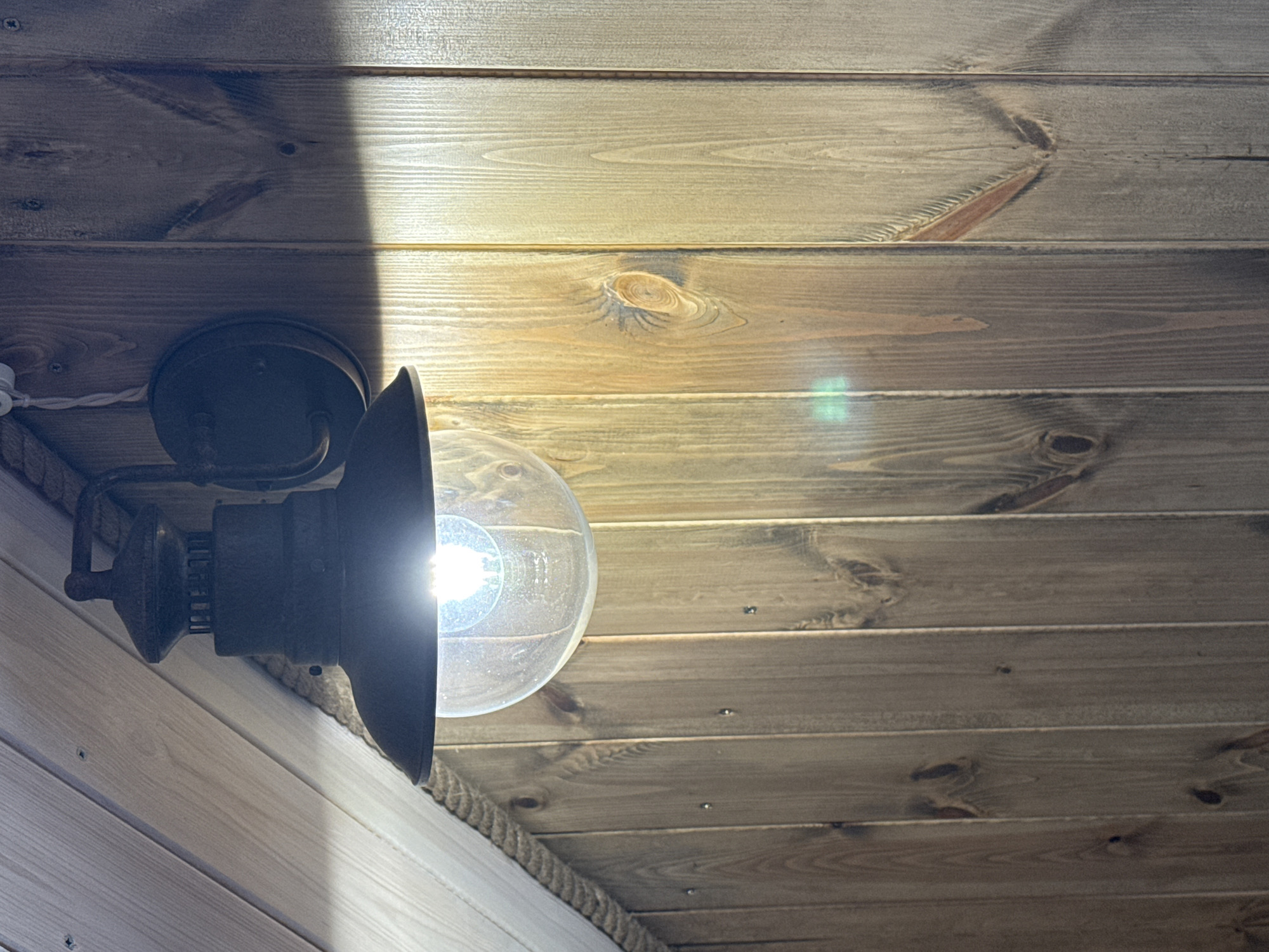 A low-angle shot reveals a room interior, possibly in Tyulyuk, Russia, given the prompt's context. The walls and ceiling are paneled with light-colored wood. A wall-mounted light fixture is attached to the left wall. The fixture has a dark-colored metal base and a clear glass globe enclosing a lit light bulb. A dark-colored shade covers the top half of the globe. The wood paneling on the wall near the light is a lighter color than the wood paneling on the ceiling. A rope trim is visible near the intersection of the wall and the ceiling. A bright, vertical line of light is visible on the left side of the ceiling.
