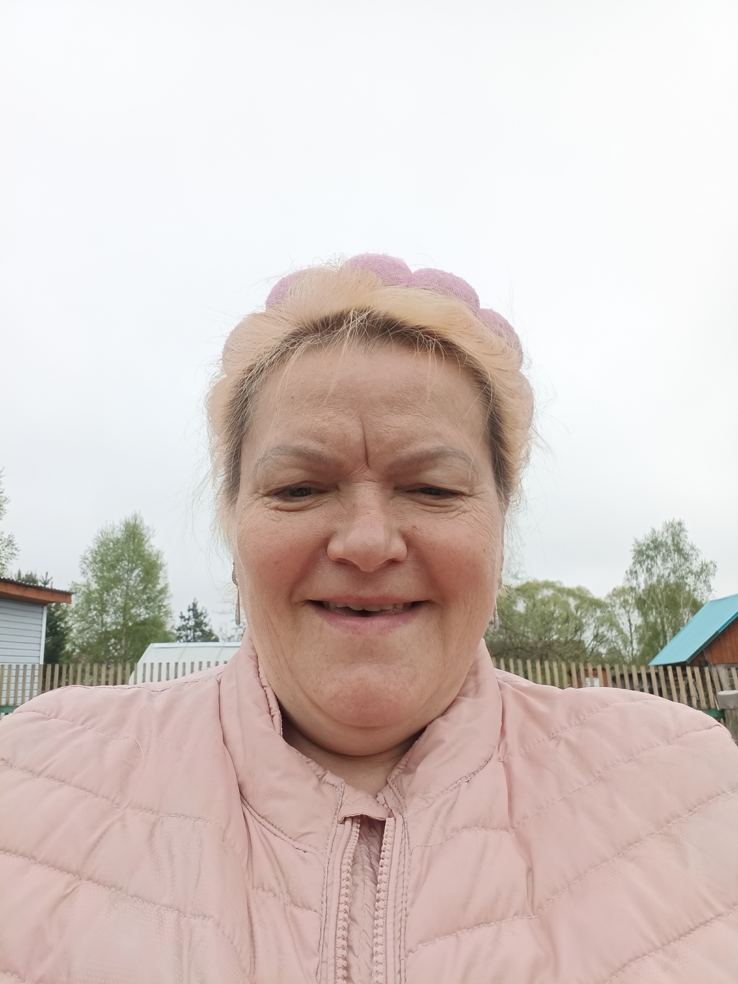A close-up, eye-level shot presents an elderly woman, her face filling most of the frame. She is smiling, and her hair, styled with pink curlers or rollers, is blonde. She wears a light pink, quilted jacket.
The background, although blurred, shows elements of an outdoor environment. Trees with green foliage are visible, along with segments of wooden fences and structures with pitched roofs painted blue. Given the prompt's mention of Красный Маяк, Russia, the scene is likely set in or near the city, suggesting a residential area possibly with gardens or small plots of land.