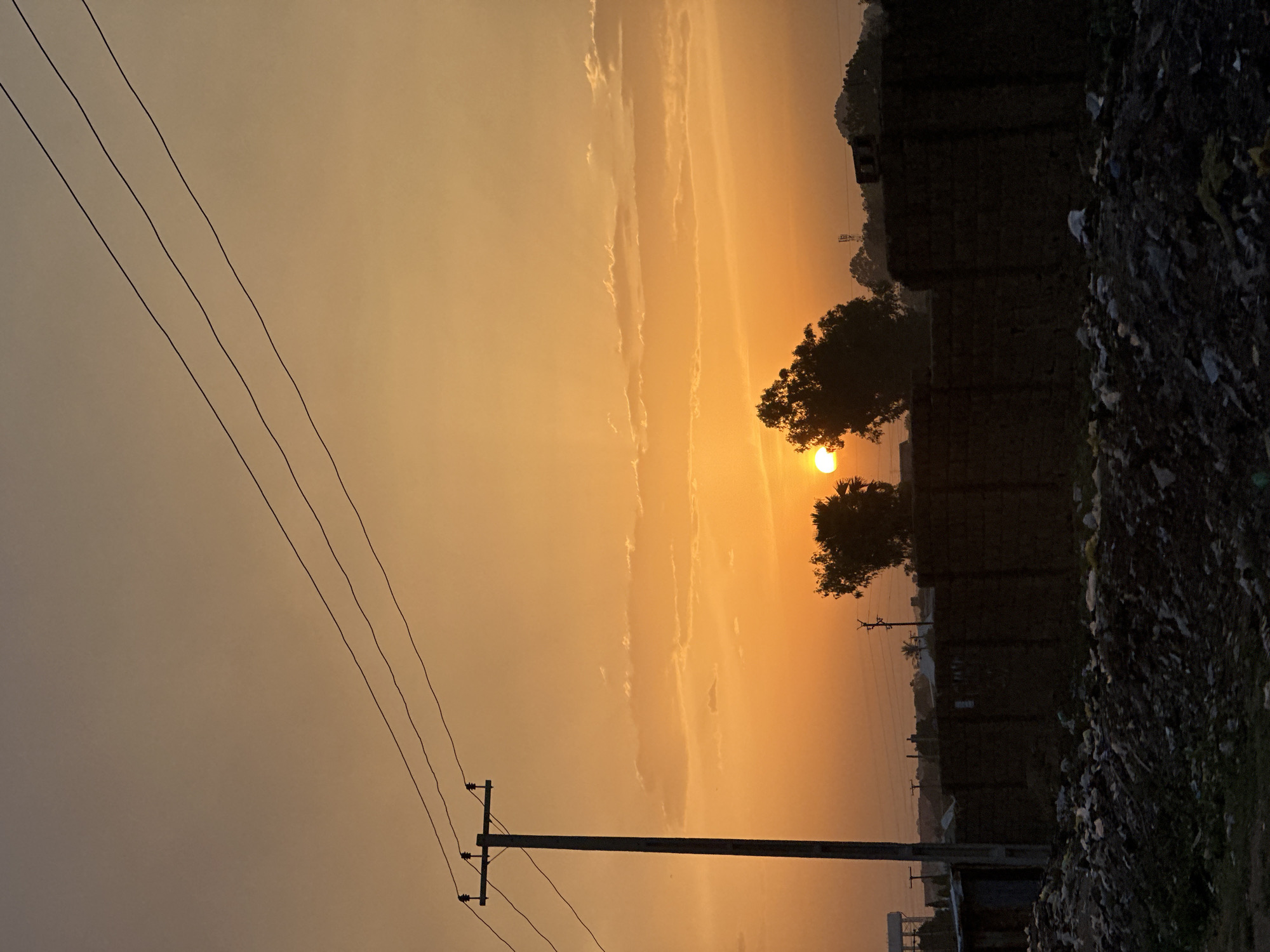 A dusk scene in Bukuru, Nigeria presents a low angle view under a hazy orange sky. The horizon shows the setting sun partially obscured by the silhouette of two trees. In the foreground, power lines run diagonally across the image, supported by a utility pole. A wall constructed of brick or similar materials spans across the middle-ground, partially obscured by piles of debris or garbage to its right. In the distance, additional utility poles and faint outlines of buildings indicate a degree of urbanization. No people are visible.