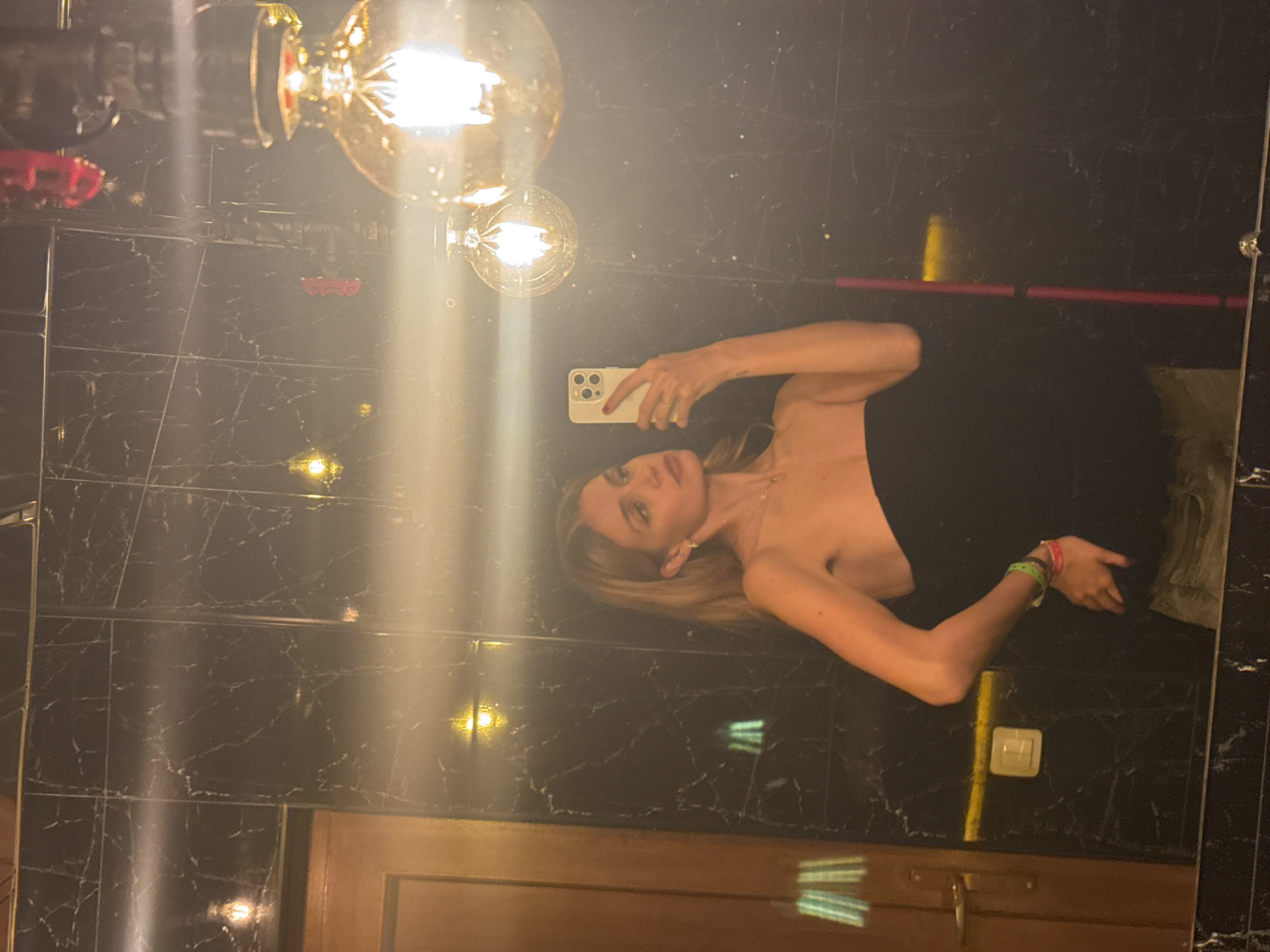 A woman is taking a selfie in a mirror in what appears to be a bathroom with black, marble-patterned tiles. The location is Yerevan, Armenia. She's holding a white smartphone, wearing a black strapless top, and several bracelets. Two Edison-style light bulbs are reflected at the top of the mirror. The reflection includes a wooden door and a light switch.