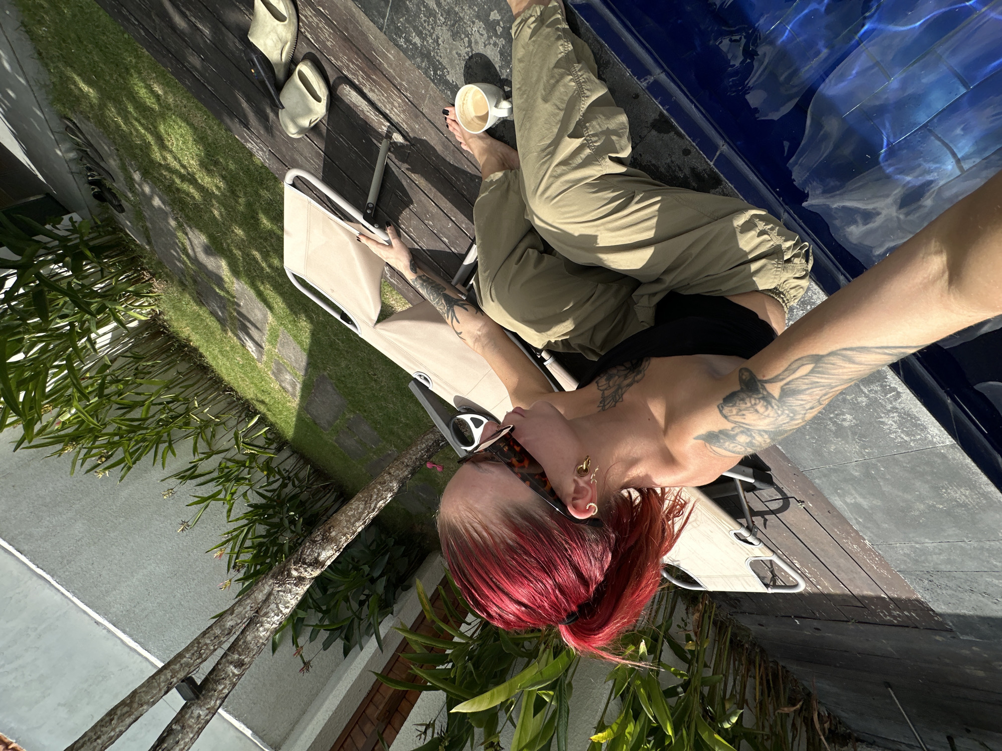 A woman with bright red hair pulled back in a ponytail and wearing sunglasses is sitting on a lounge chair next to a swimming pool. She is wearing a black top and olive green pants. She is holding a cup of coffee in her right hand. The lounge chair is beige and is on a wooden deck. There are plants and a wall in the background. The swimming pool is blue and appears to be outdoors. The image was taken in North Kuta, Indonesia.