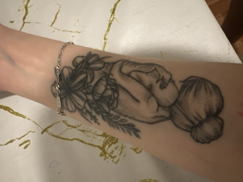 This image, taken in Chaikovsky, Russia, presents a close-up view of a fair-skinned human forearm. The arm is positioned diagonally across the frame, resting on a white or off-white surface exhibiting an irregular, sparse pattern of thin gold-yellow lines, consistent with a veined material or staining.
A monochrome tattoo is prominently displayed on the inner aspect of the forearm. The tattoo depicts two adult hands in a cradling posture, holding an infant whose head and upper torso are visible. Above the hands, a dense cluster of various flowers and foliage is tattooed, resembling a bouquet. Below the hands, a grouping of approximately three textured objects, consistent with walnuts or similar nuts, is rendered.
Near the wrist, a delicate, silver-colored chain bracelet is worn. The bracelet features a small, rectangular metallic charm and another small, decorative element, possibly a star or cross, attached to the chain.