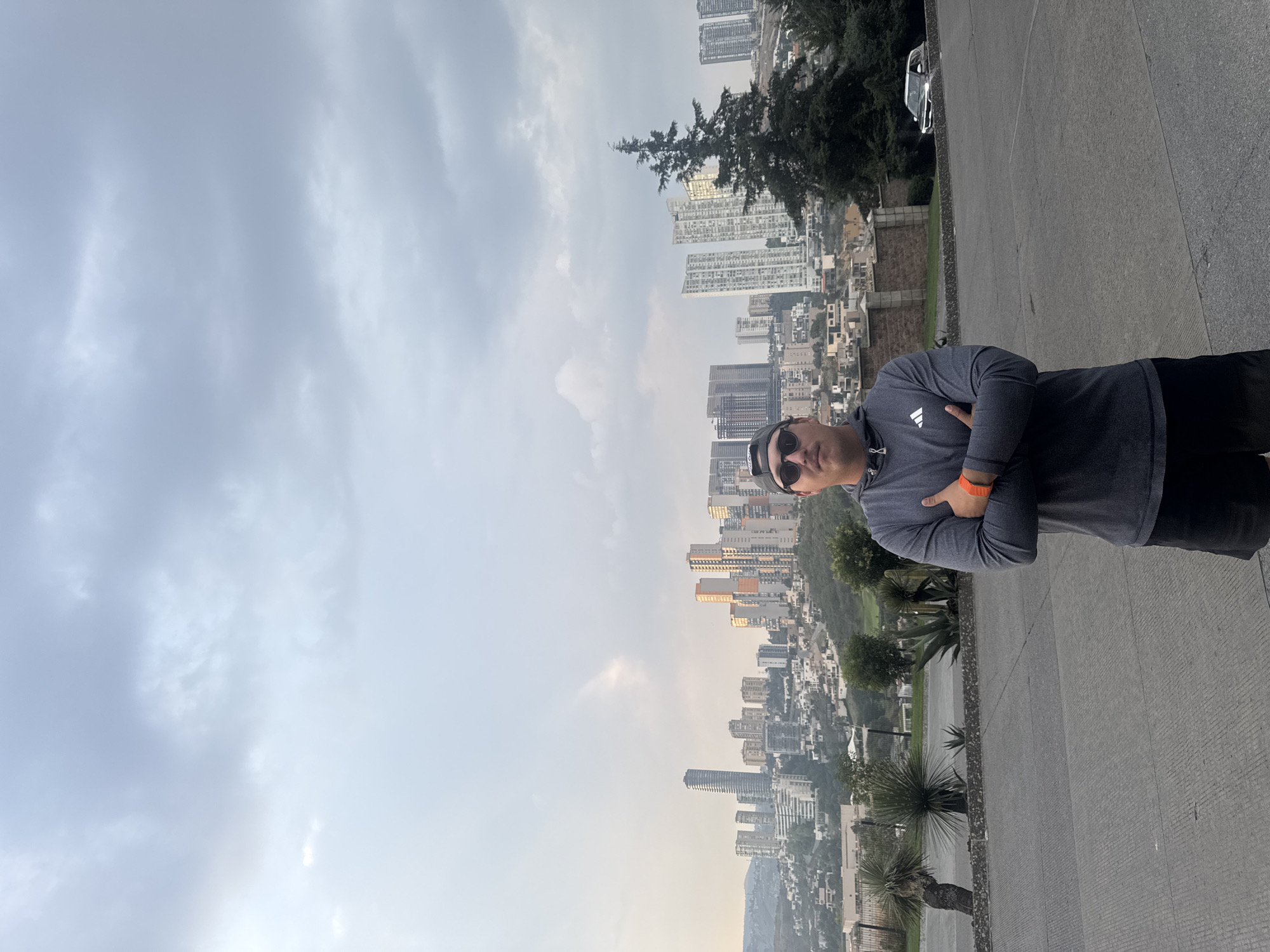 A medium shot shows a man in sunglasses, a long-sleeve athletic shirt with an Adidas logo, shorts, and an orange bracelet, standing with arms crossed, on a paved walkway in the city of Huixquilucan de Degollado, Mexico. Behind him, across the walkway, is a cityscape featuring a cluster of tall, modern apartment buildings and office towers, with green trees and some lower structures visible in between. A line of trees stands above the buildings. The sky is a mix of grey and white, suggesting overcast or hazy conditions. In the top right corner, a parked silver car is visible.
