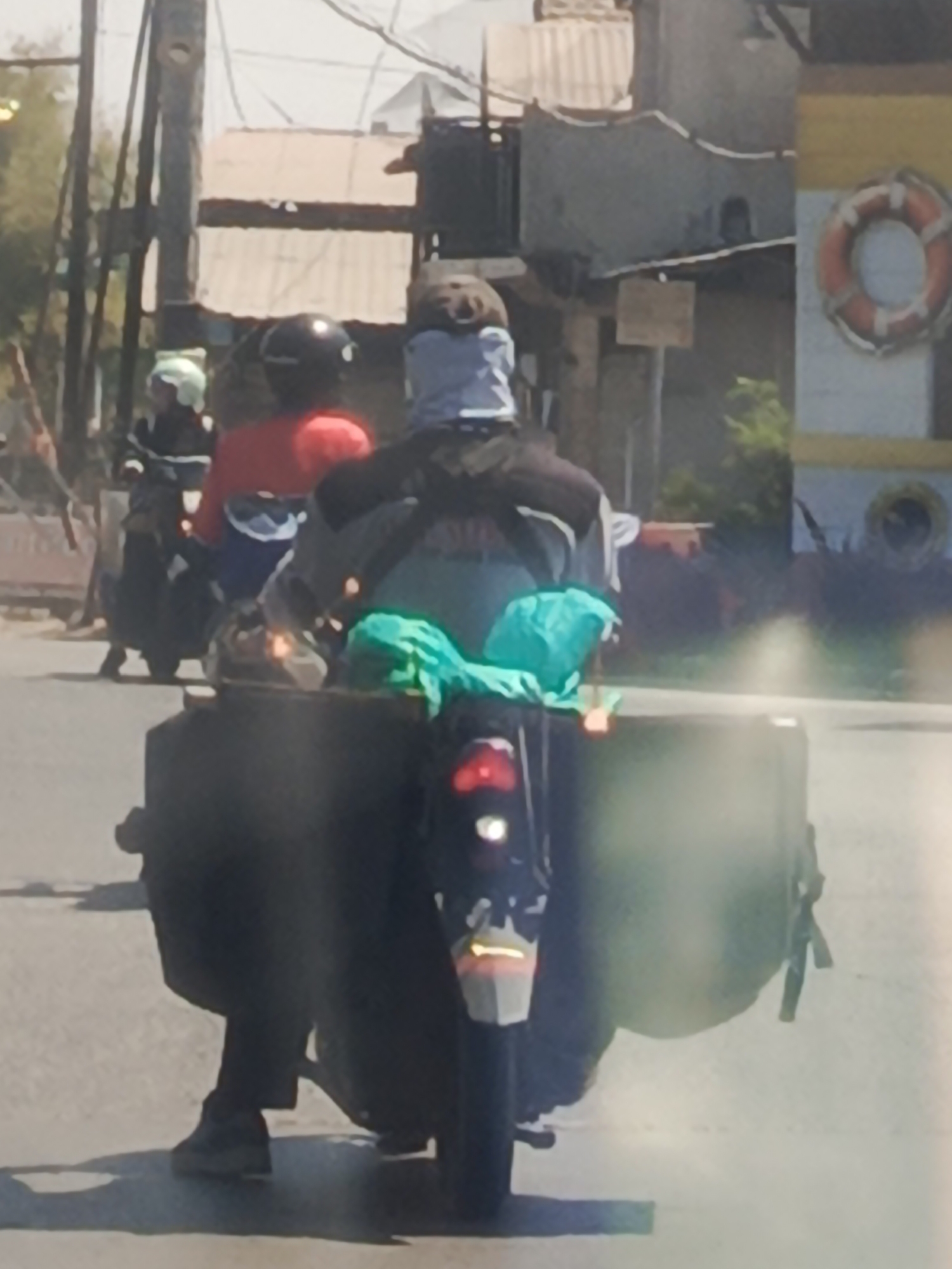 A blurry, medium-distance shot shows a person riding a motorcycle on a street in Pasinan Lemahputih, Indonesia. The person is wearing a grey head covering and a grey and black jacket, and appears to be carrying a large, dark-colored container on the back of the motorcycle, possibly a cooler or a cargo box. Two bright green bags are tied to the top of this container. The motorcycle has a red taillight. In the background, there are buildings with corrugated metal roofs and some foliage. A life preserver is visible on the side of a structure on the right.