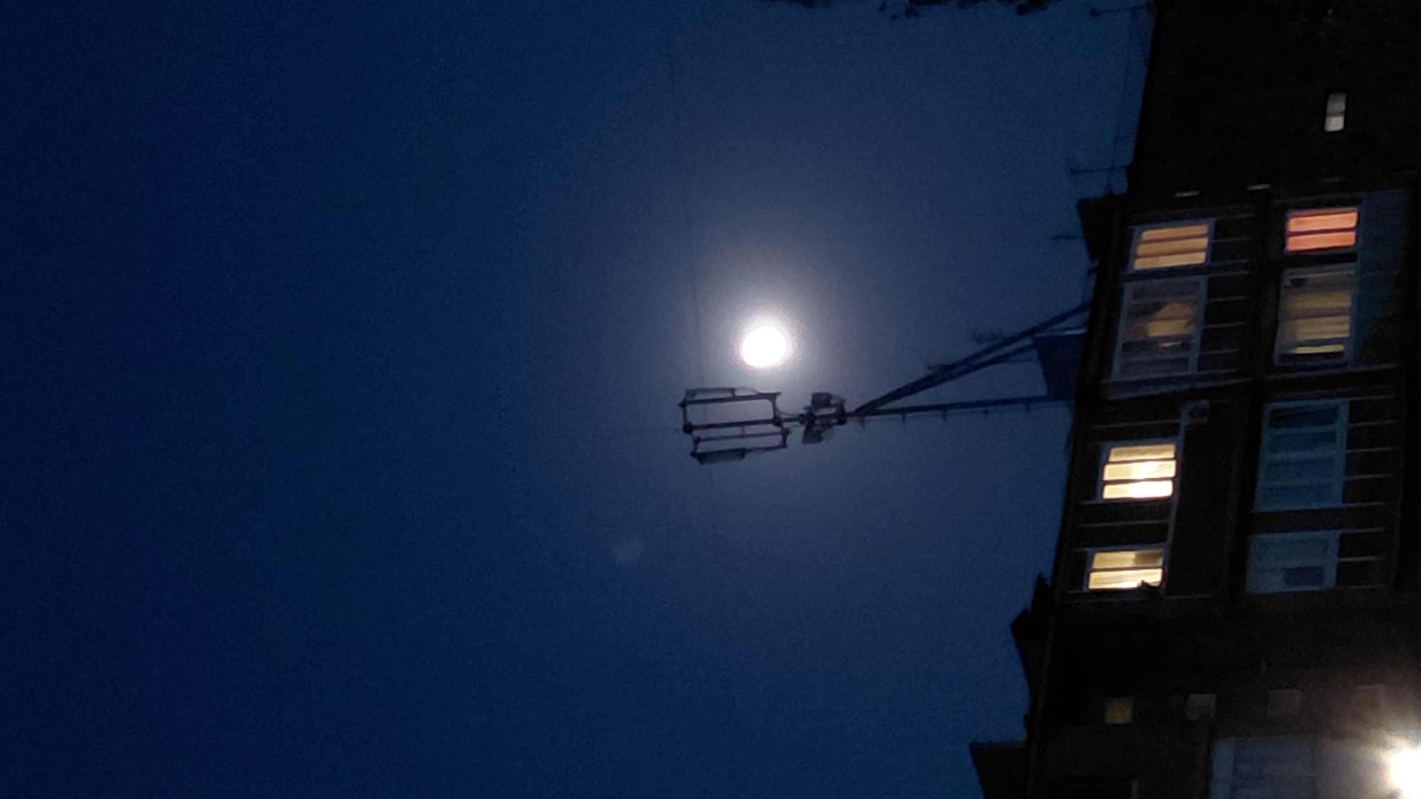 A dark blue night sky is visible above a residential building in Abovyan, Armenia. A cellular tower, equipped with antennas, is silhouetted against the sky. A bright, full moon is positioned to the right of the tower. The building's facade is dark, with several windows illuminated from within. Some windows display warm yellow and orange light, while others show a cooler, possibly blue or white light. The overall scene depicts a nighttime urban landscape.