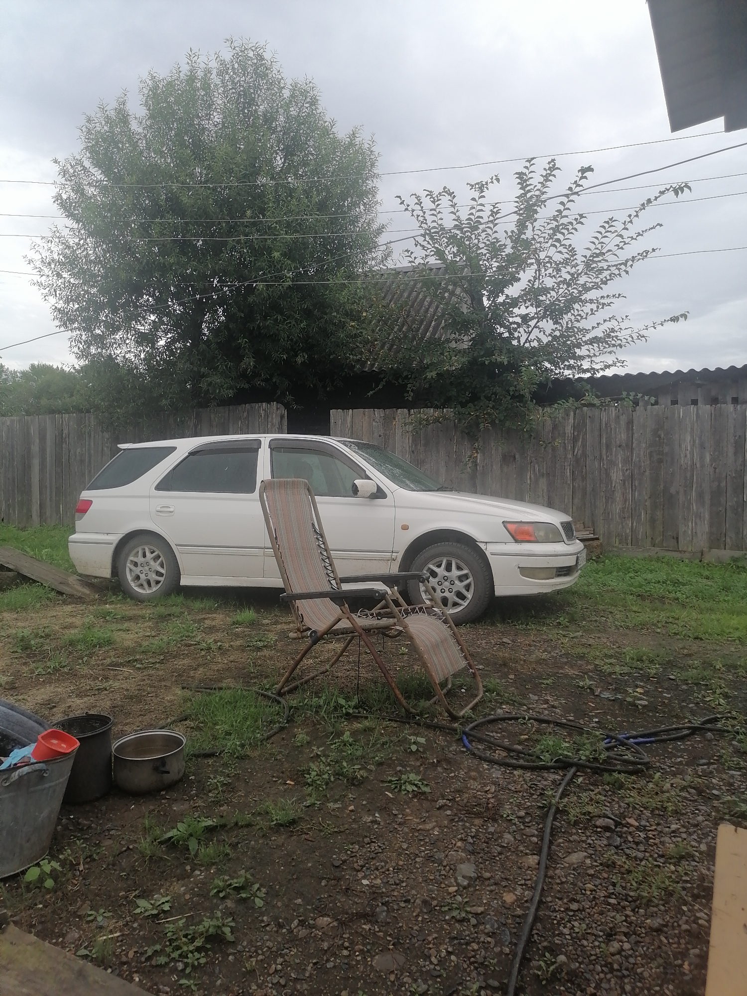 A white station wagon is parked on a grassy and gravelly area next to a wooden fence. A folding lawn chair is positioned in the foreground. Several buckets and a large pot are to the left of the car. A hose lies on the ground near the chair. Trees and overcast skies are visible in the background. This scene is likely in Novopokrovka, Russia, given the style of the house and fence.